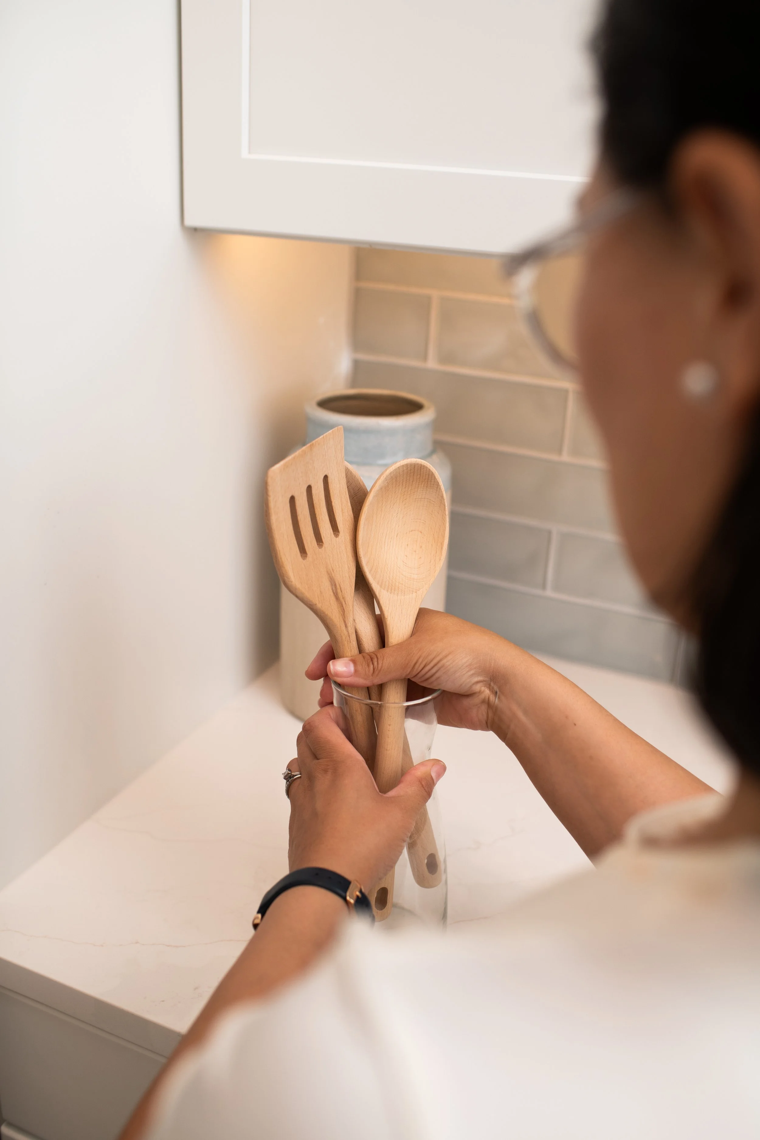 A person holding wooden kitchen utensils including a spatula and a spoon in a glass on a white countertop in a kitchen.