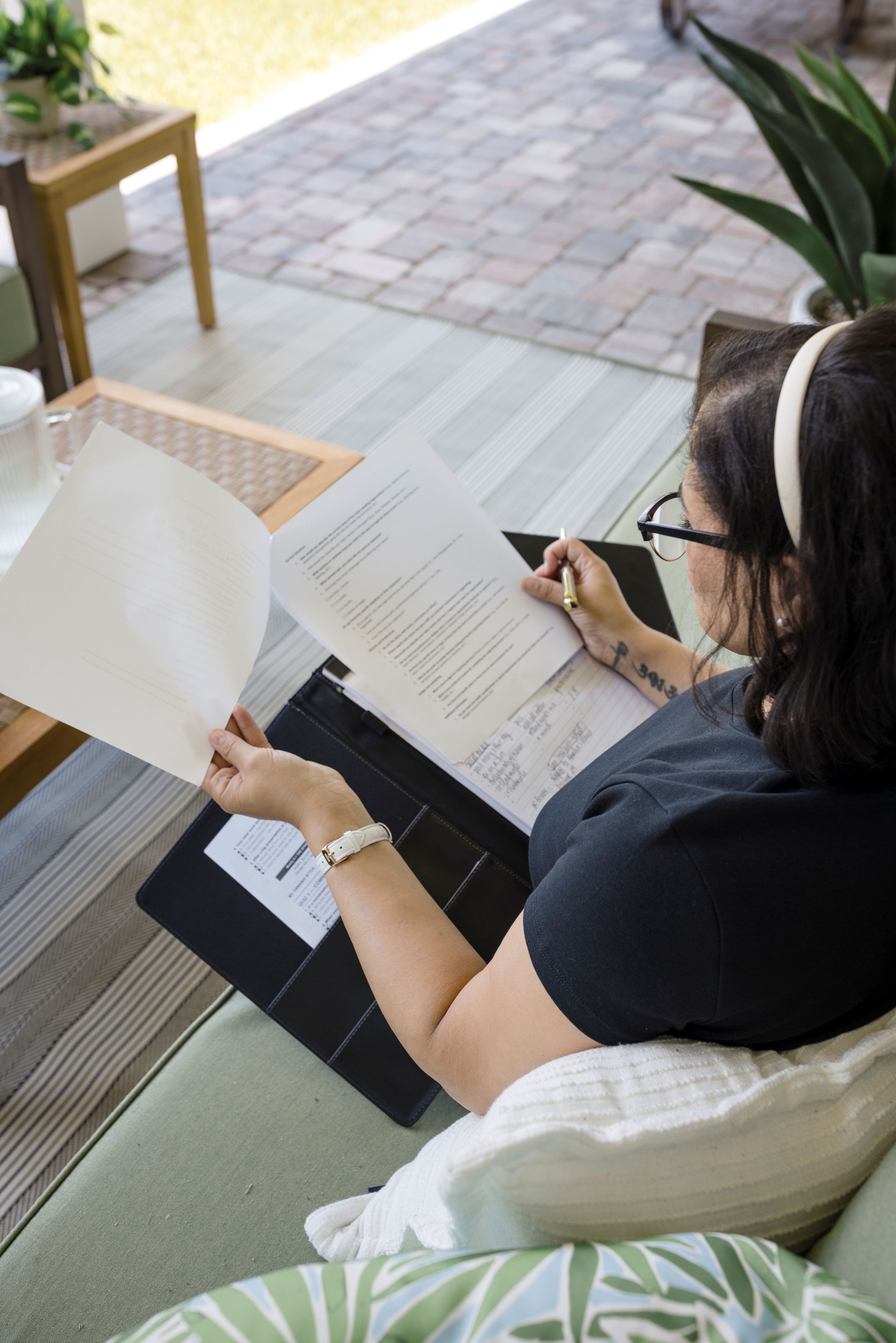 Woman with dark hair and glasses sitting on a patio, reading papers, with plants and outdoor furniture nearby.