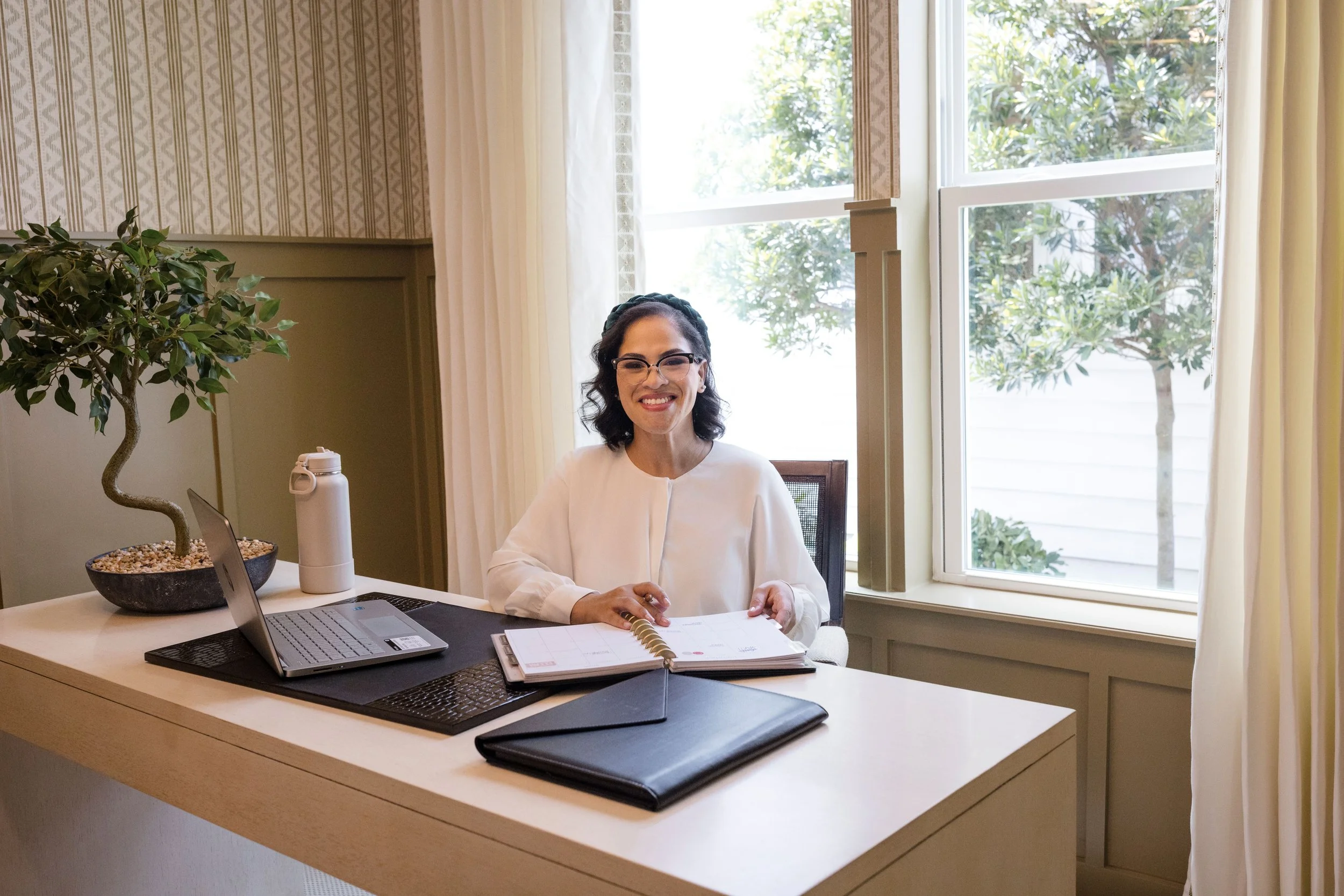 A woman with glasses is sitting at a desk with a laptop, notebooks, a water bottle, and a potted plant, smiling at the camera in a room with large windows and curtains