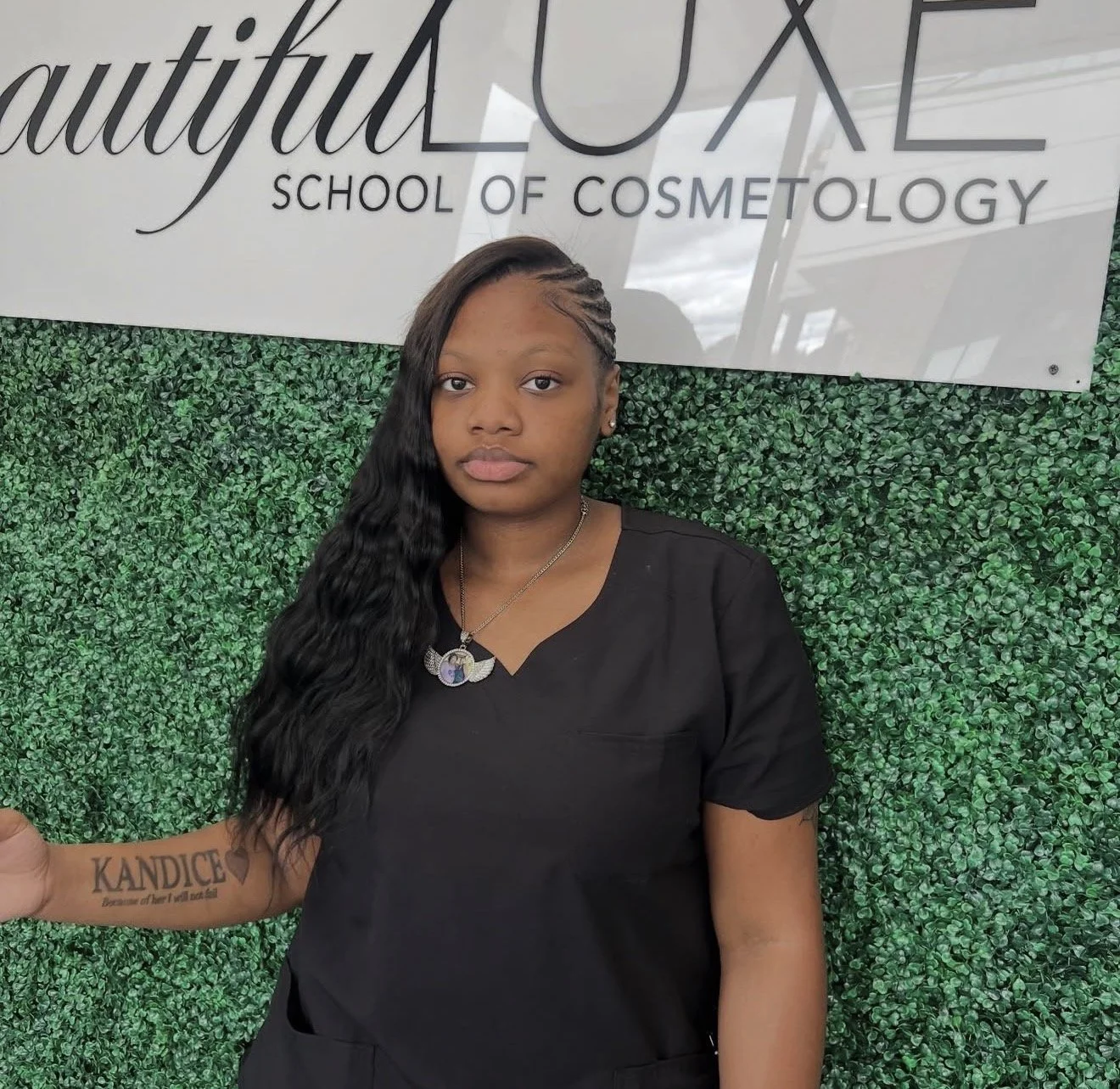CTE Cosmetology student standing in front of the Beautiful Luxe School of Cosmetology sign, wearing black scrubs and showcasing a finished hairstyle in a professional setting