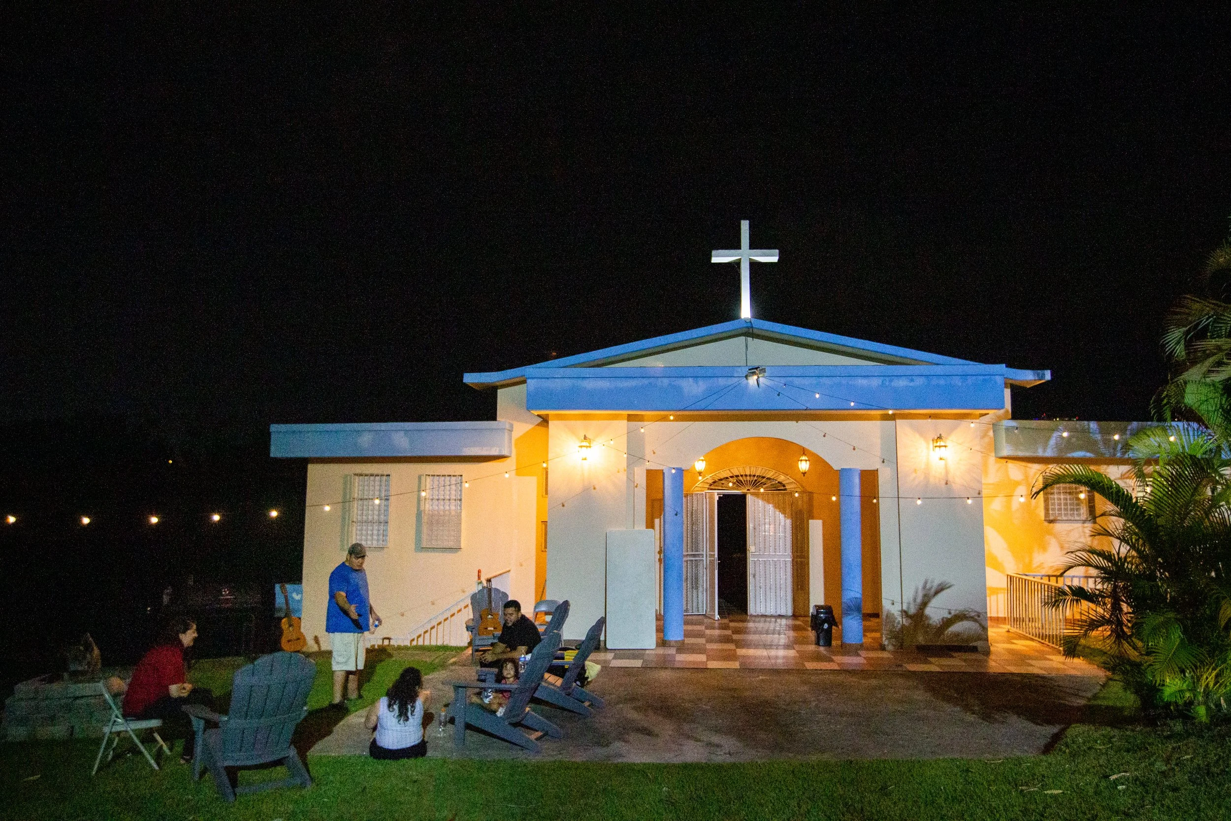 Iglesia cristiana con cruz en la parte superior, iluminada en la noche, con personas sentadas en el pasto y en sillas, y guitarras apoyadas contra la iglesia.