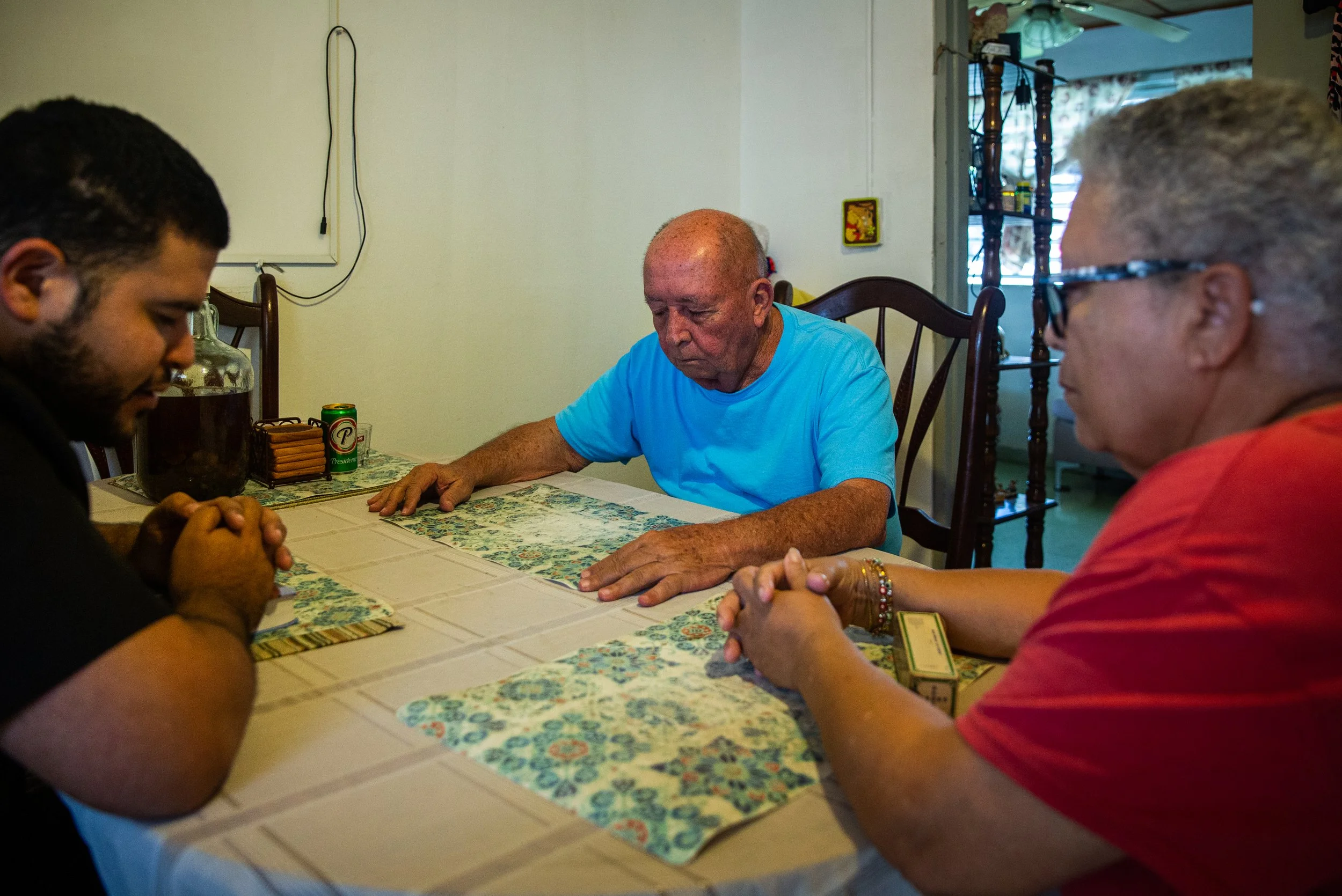 Tres personas mayores y un joven orando en la mesa de la sala, con las manos juntas y la cabeza inclinada, en una postura de oración o reflexión.