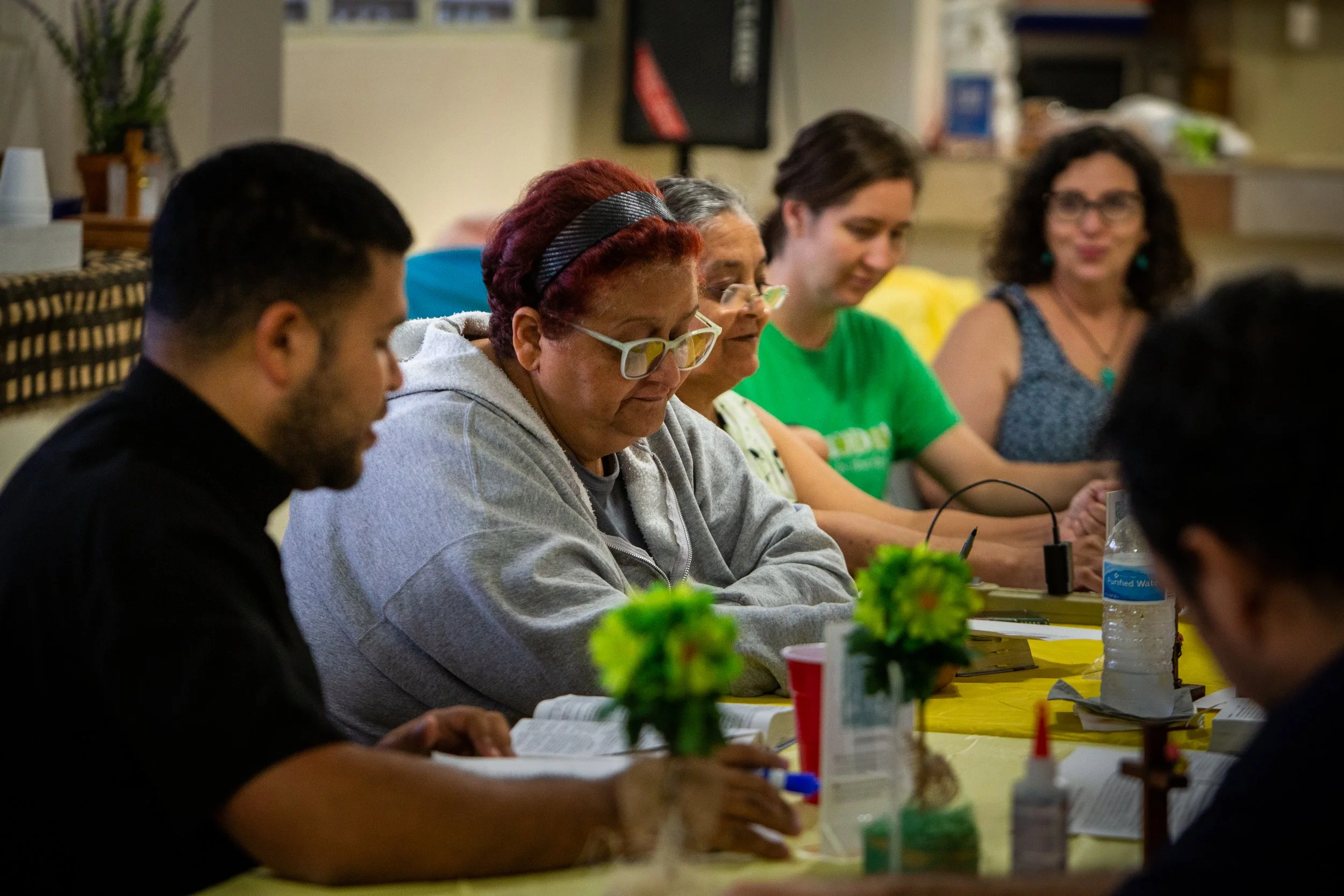 Grupo de seis personas sentadas en una mesa de comedor, leyendo o estudiando, con libros y objetos como botellas de agua y adornos en el centro de la mesa.