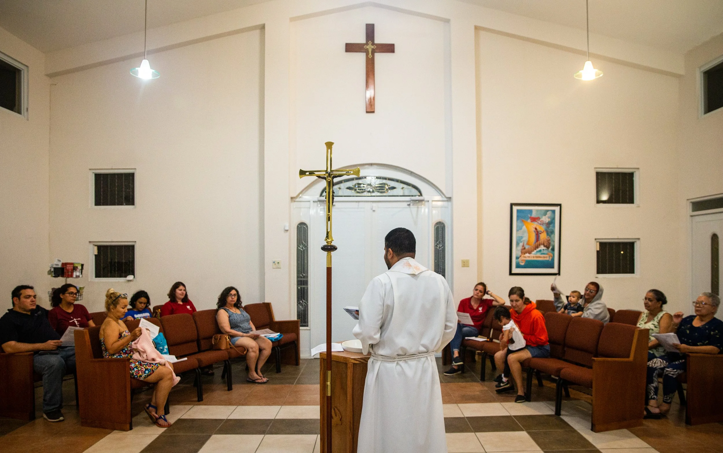 Un pastor en una iglesia con un grupo de personas sentadas en bancos de madera, en el fondo hay un crucifijo grande en la pared y un cuadro con una nave y una montaña, durante una ceremonia religiosa.