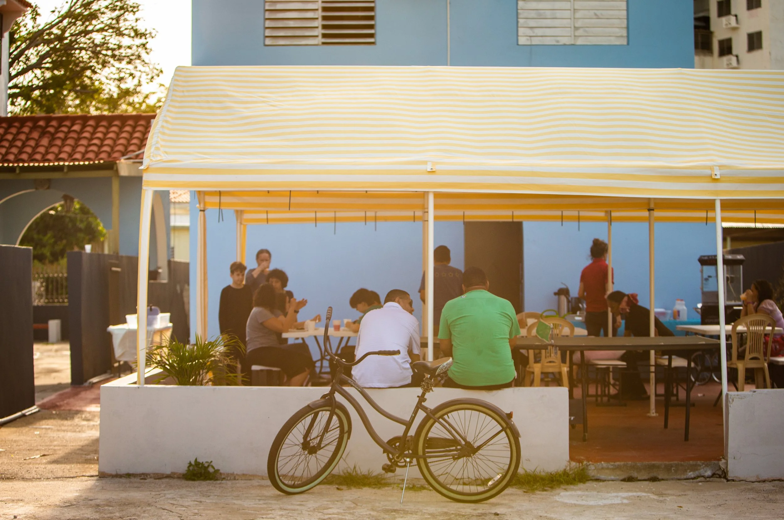 Grupo de personas sentado en mesas bajo una carpa amarilla con rayas blancas, en un espacio abierto con edificio azul al fondo, y una bicicleta en primer plano.