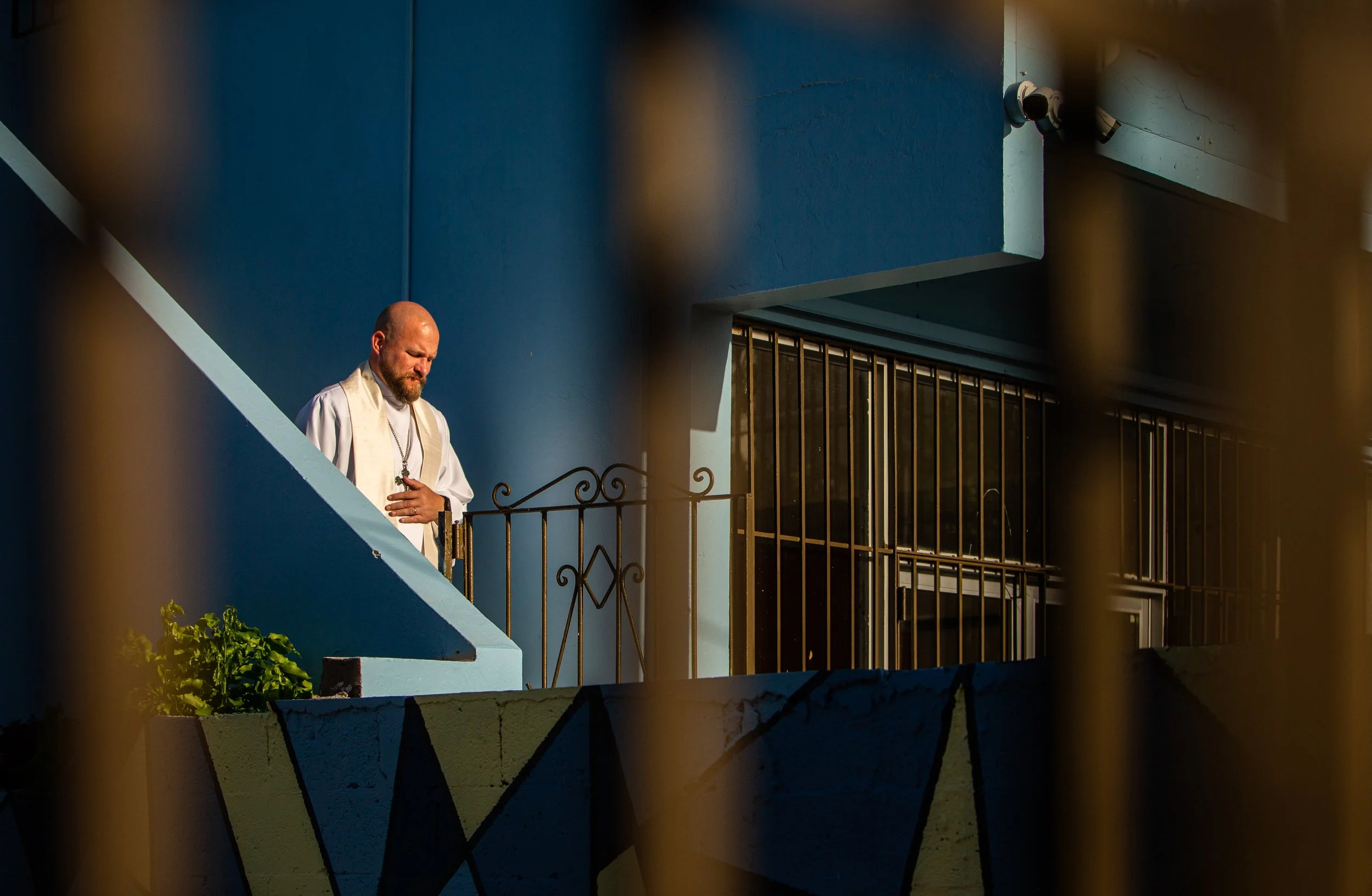 Hombre con vestimenta religiosa haciendo oración en un balcón, visto a través de una reja metálica, con pared azul de fondo y barandillas negras.