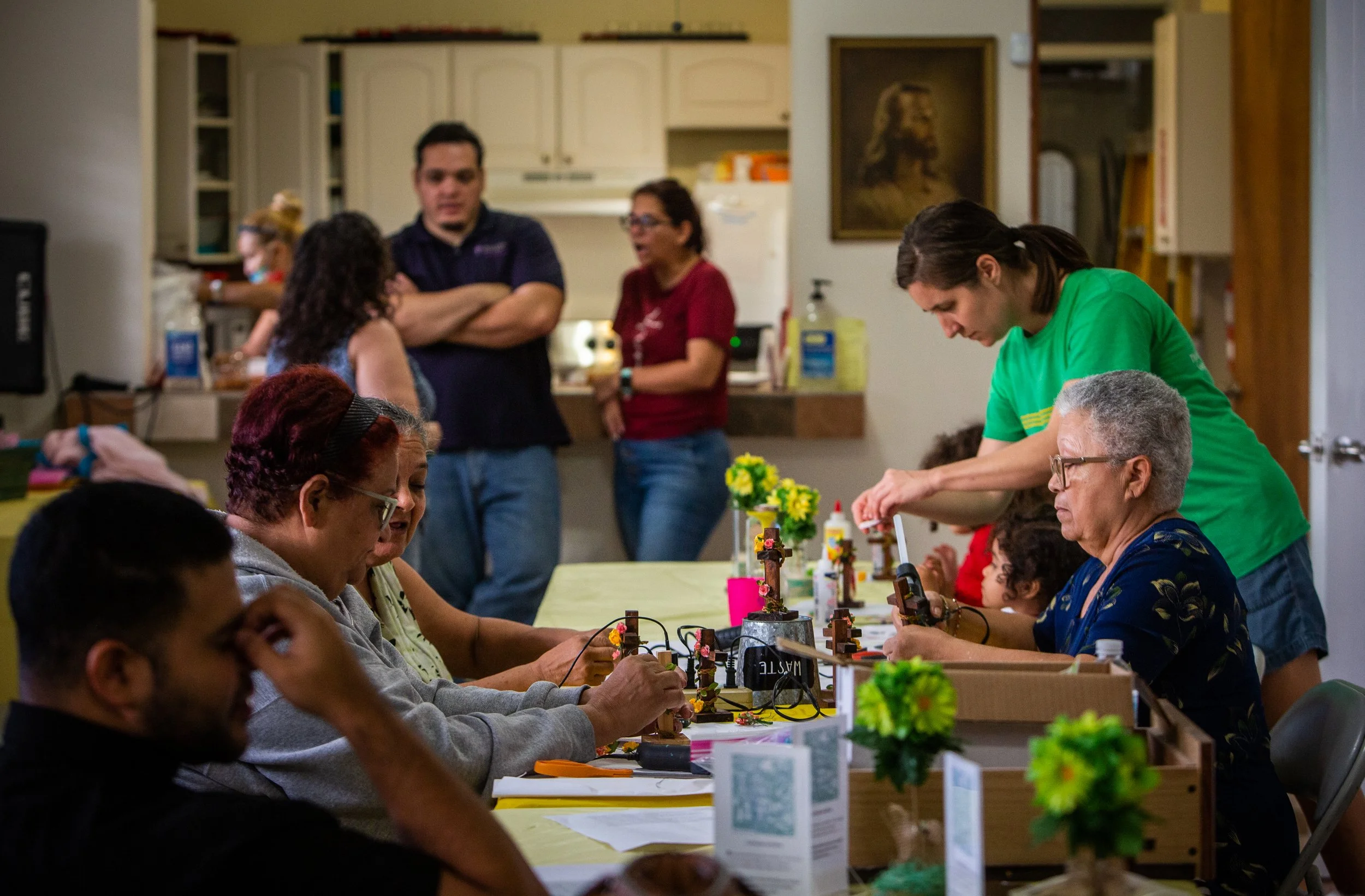 Grupo de personas participando en un taller de artesanías en una mesa con decoraciones y materiales de manualidades, mientras otros conversan y observan en el fondo.