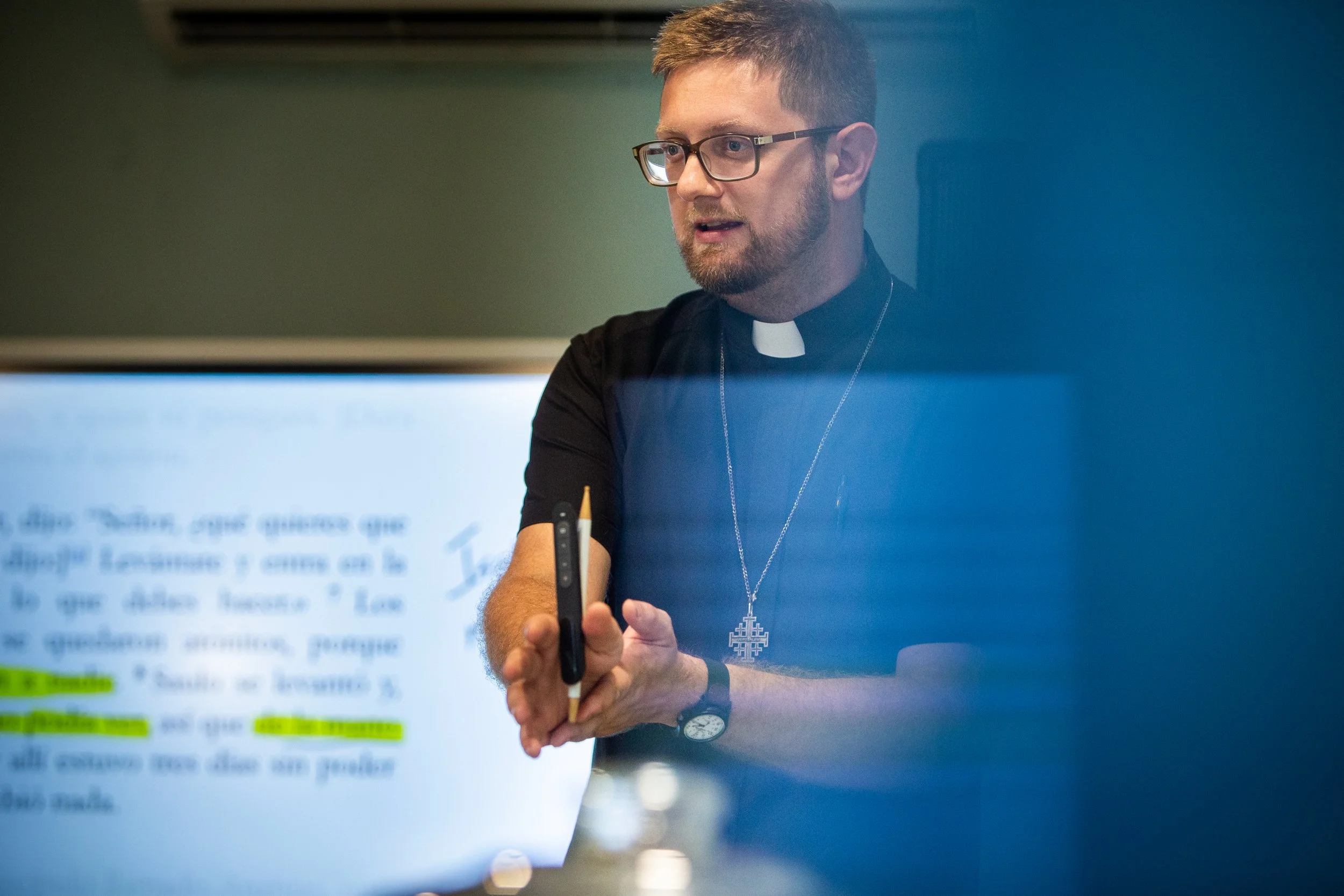 El Pastor James Neuendorf con gafas, barba y una cruz en el cuello, sosteniendo un bolígrafo y dando una explicación en una pizarra digital con texto en español.