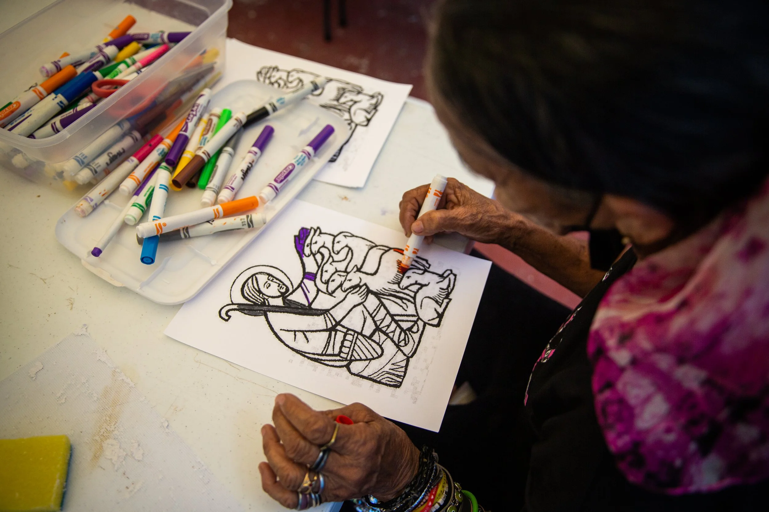 Anciana coloreando un dibujo de una figura femenina vestida con ropa tradicional usando marcadores en una mesa