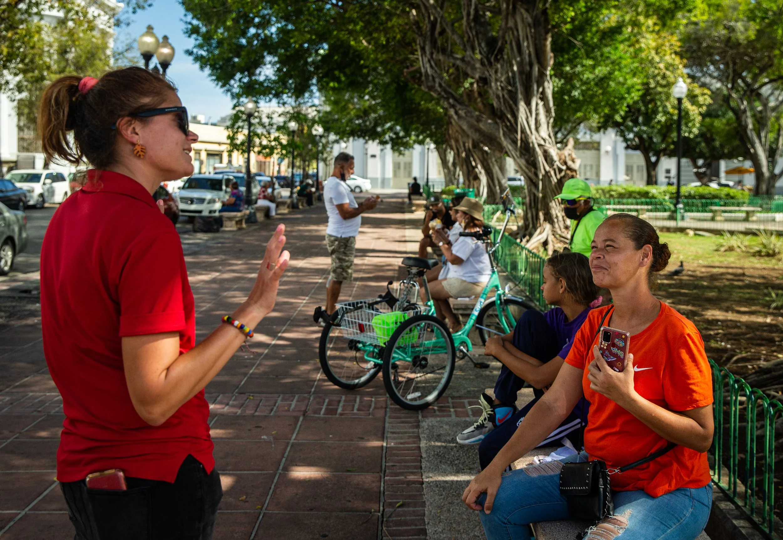 Mujer de cabello rubio con gafas negras, camiseta roja y pulsera, hablando con un grupo de personas en un parque. Algunas personas están sentadas en bancos, usando mascarillas, y otras de pie, algunas usando bicicletas y sombreros.