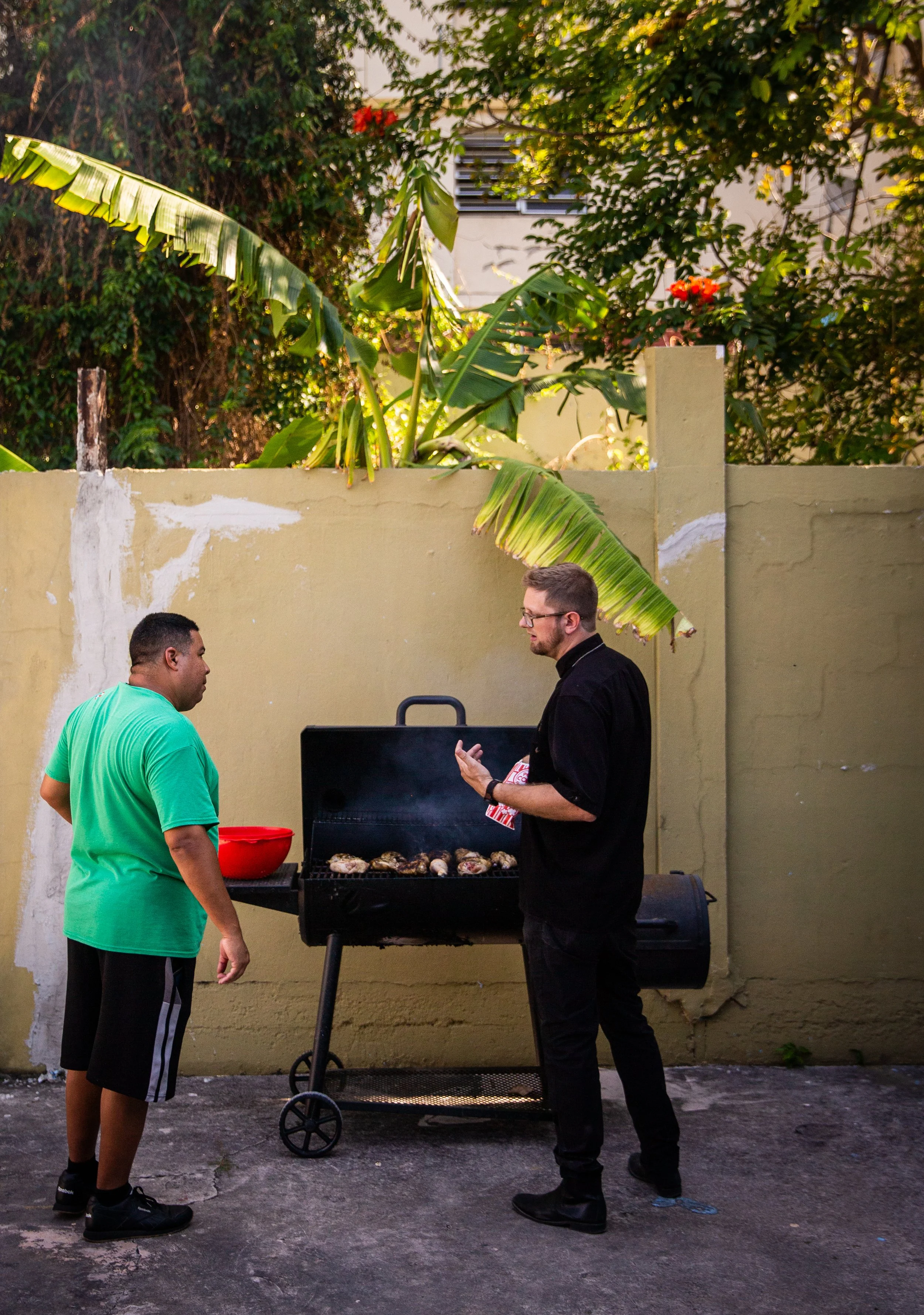 Dos hombres conversando junto a una parrilla de barbacoa en un patio con muro de fondo, uno de ellos está cocinando carne y el otro le habla mientras sostiene un paquete, en un ambiente exterior con vegetación y un muro de color beige.