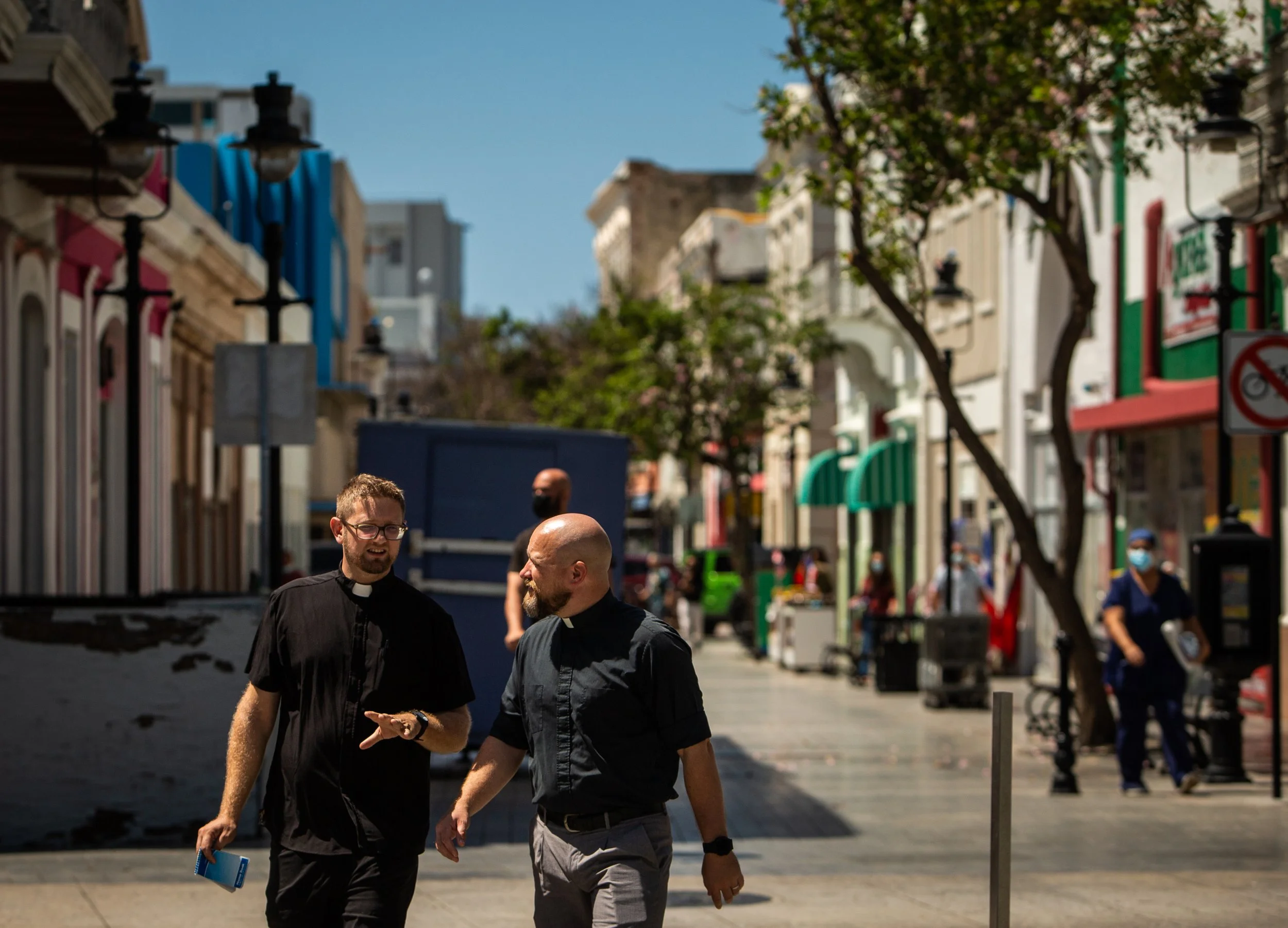 Dos pastores caminando y conversando en una calle peatonal con tiendas y árboles, algunos personas con mascarillas en el fondo.