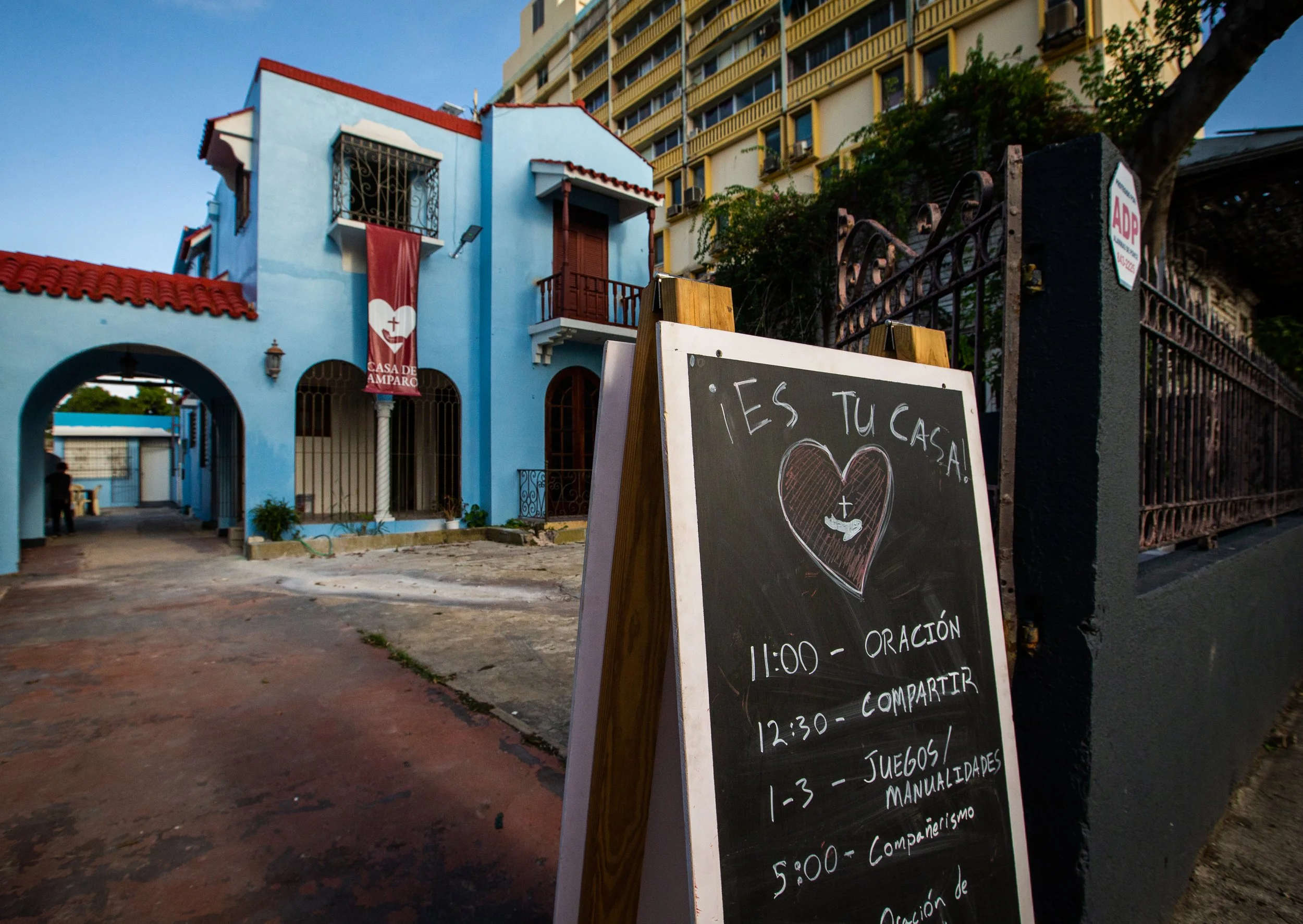 Cartel en la calle, con horarios y actividades para un evento en una casa de misericordia, frente a una casa de azul con rejas y un árbol, en un entorno urbano.