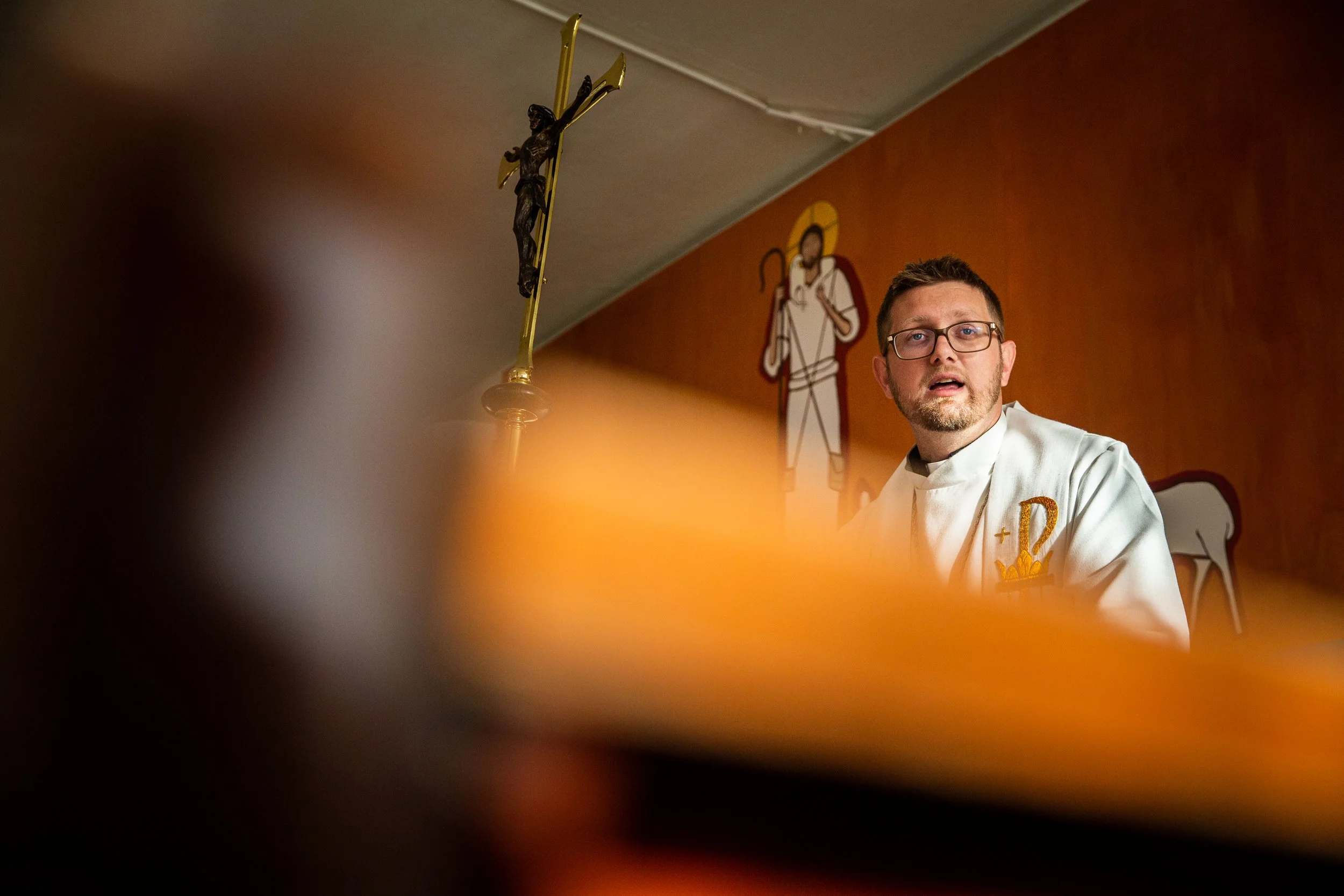 Un pastor con gafas en una iglesia, con una cruz y un dibujo religioso en la pared de fondo.