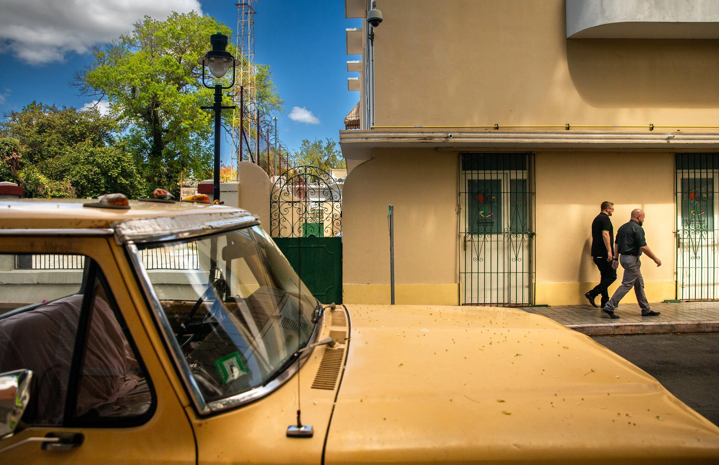 Una calle con un coche amarillo en primer plano, y en el fondo un edificio beige con rejas negras en las ventanas. Dos hombres caminan por la acera, uno con camiseta negra y pantalón oscuro, y otro con camisa negra y pantalón gris. Hay árboles verdes