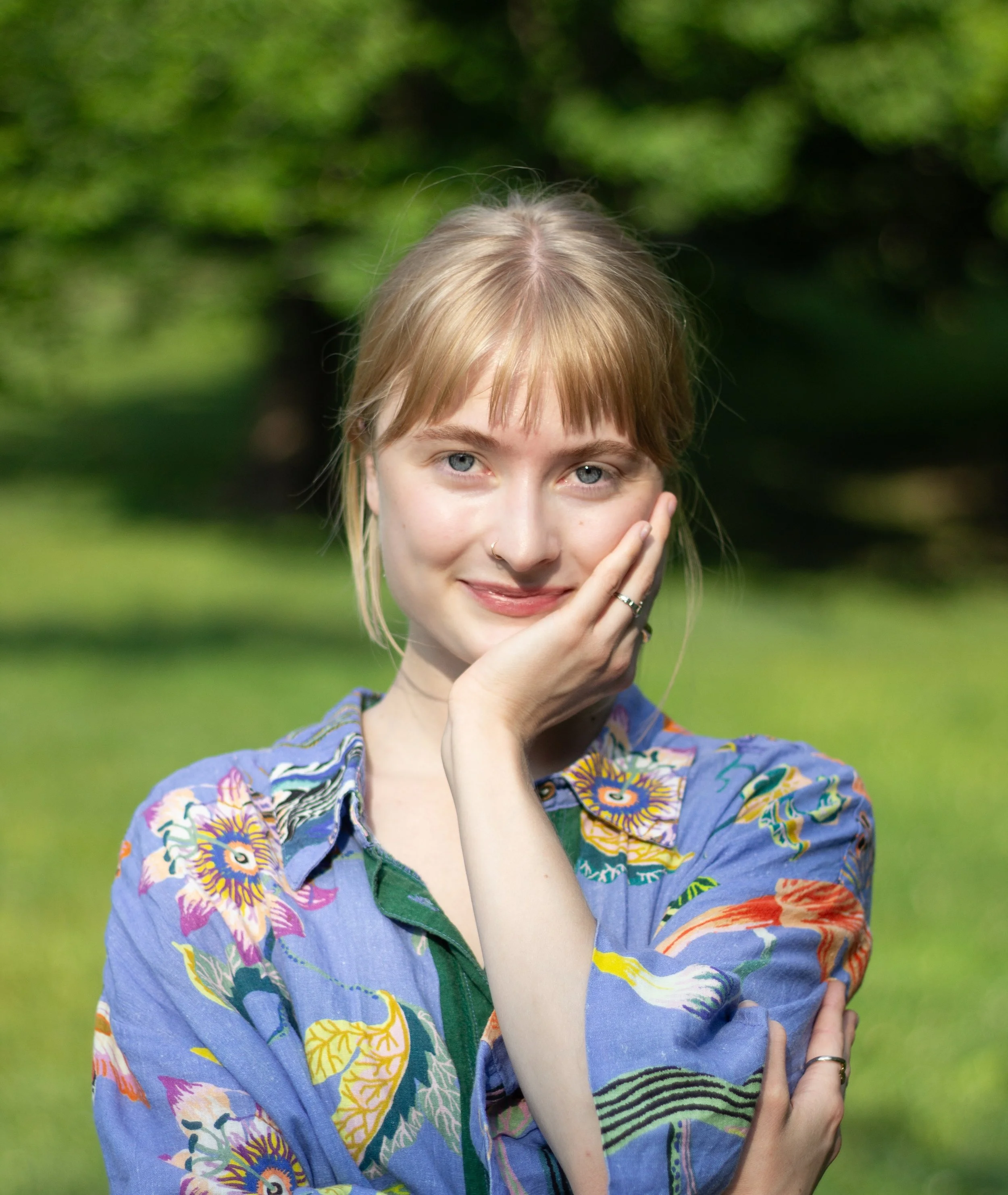 A close-up image of Maria smiling gently with their left hand resting on their left cheek. She is wearing silver rings. Her hair is tied back with wisps falling out. They are wearing a bright blue collared shirt that is a floral tropical pattern.