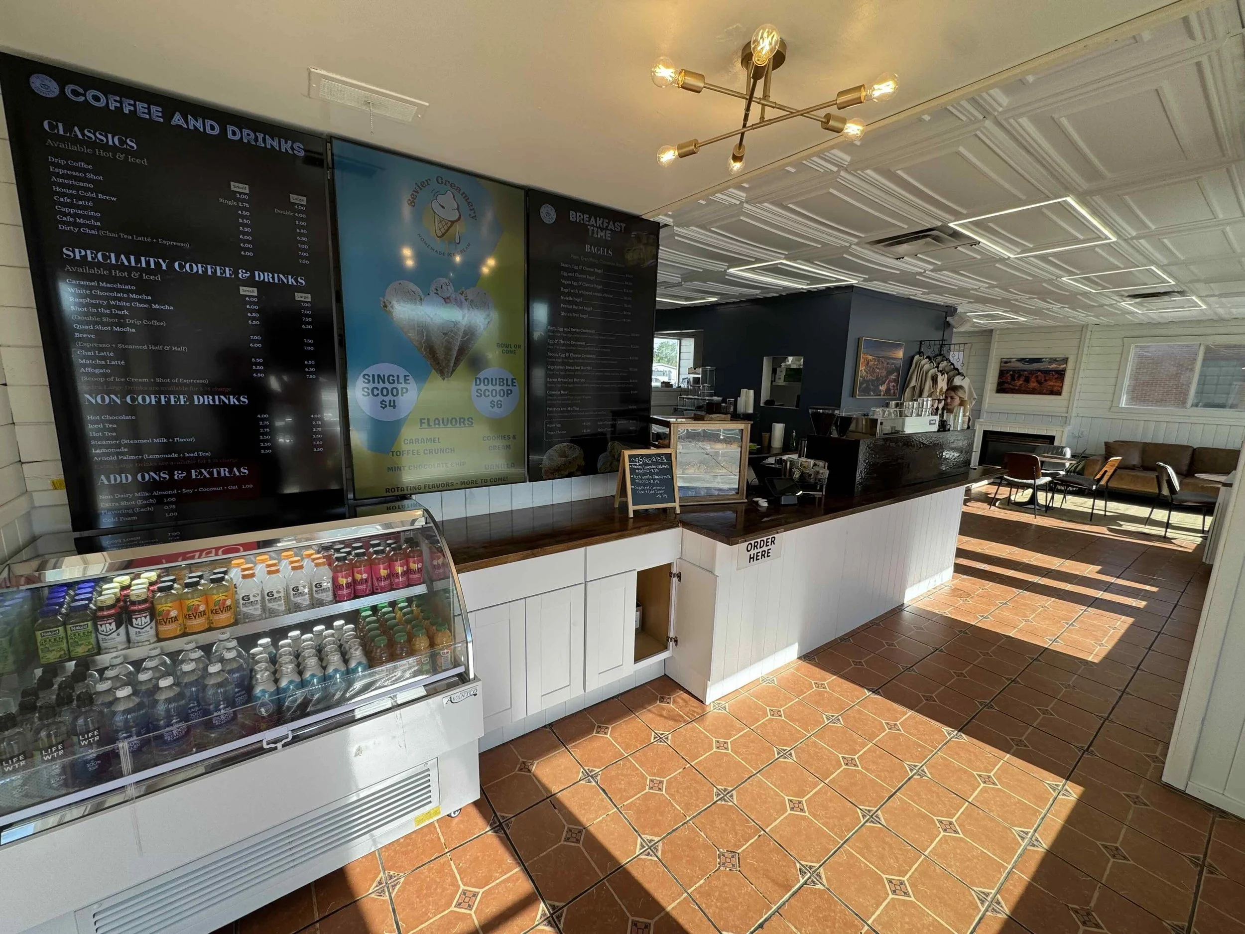 Interior of a coffee shop with black menus on the wall listing coffee drinks, a refrigerated display with bottled beverages, and a counter for placing orders, with sunlight streaming through windows and artwork on the walls.