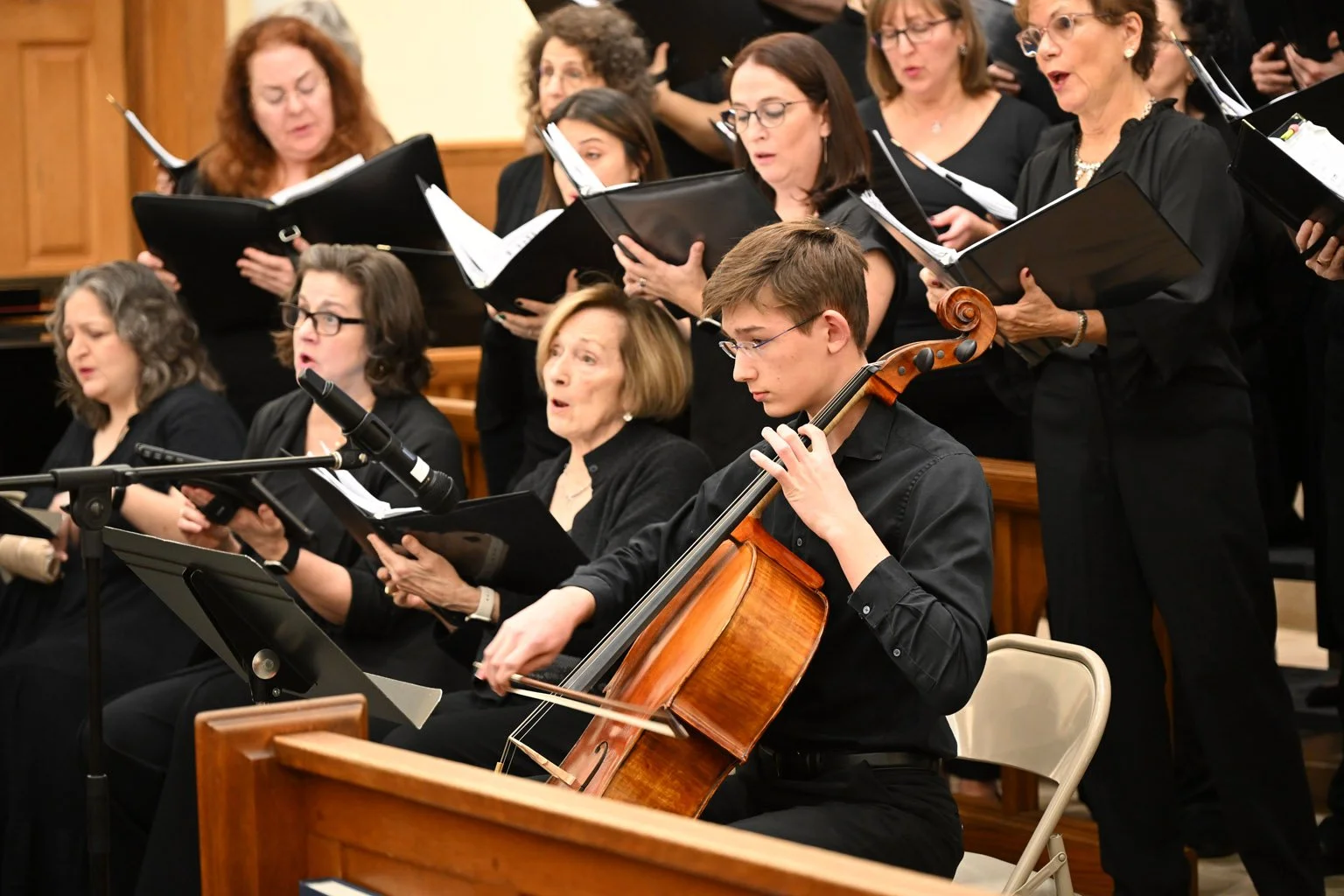 Photo of a group of women all dressed in black, singing, with folders held in their hands and in the foreground, a young, caucasian boy plays the cello