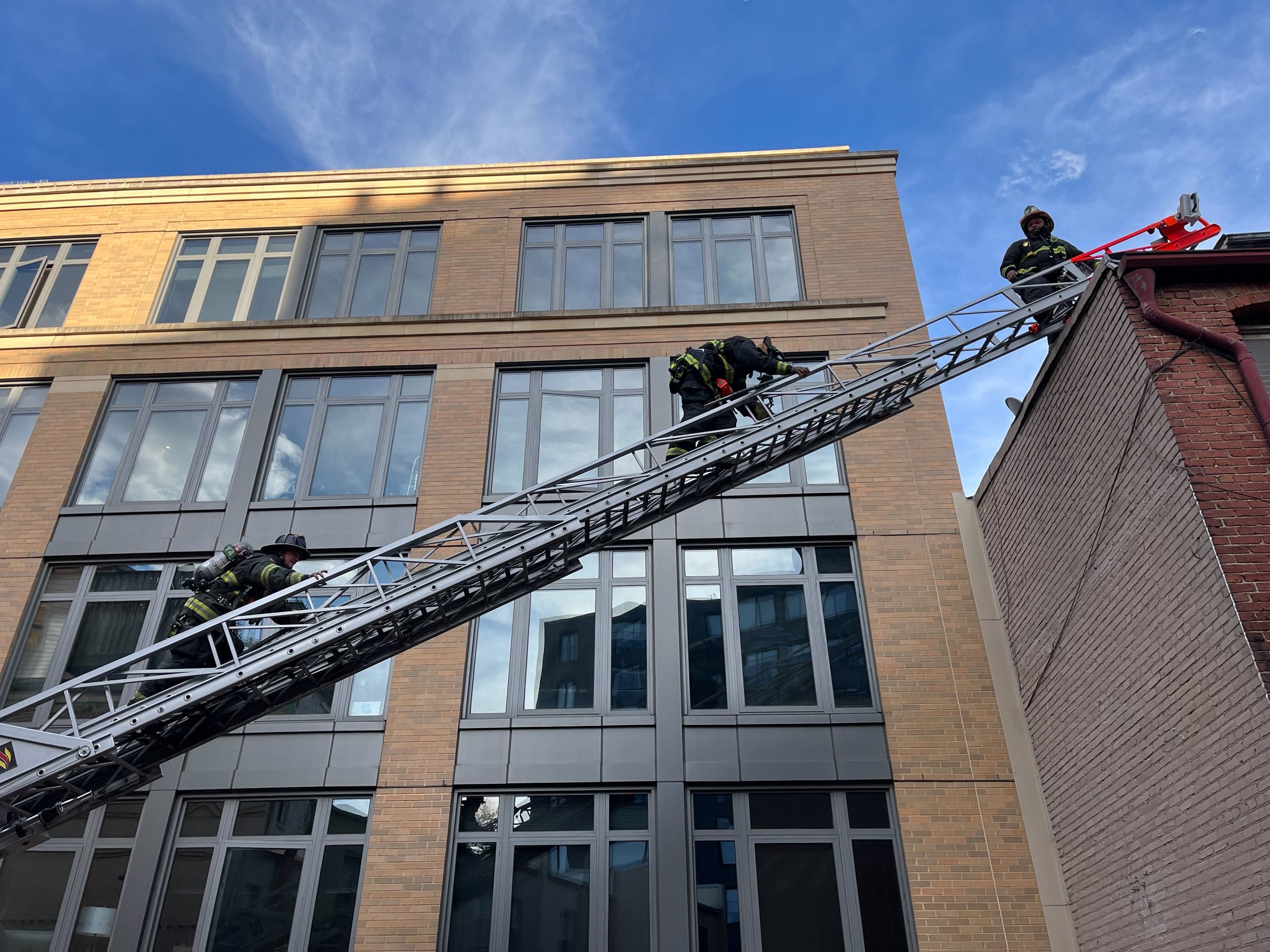 Firefighters climbing a ladder with a large building in the background.