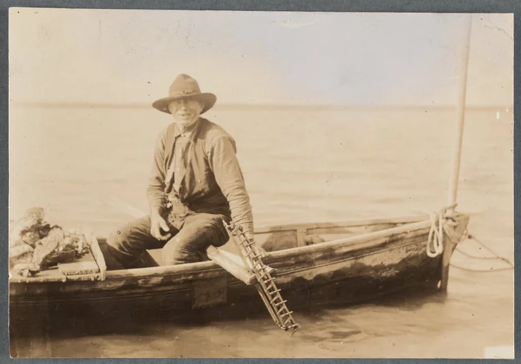 An oysterman sits in a boat with his tongs and other oystering tools. New York Public Library Digital Collections.