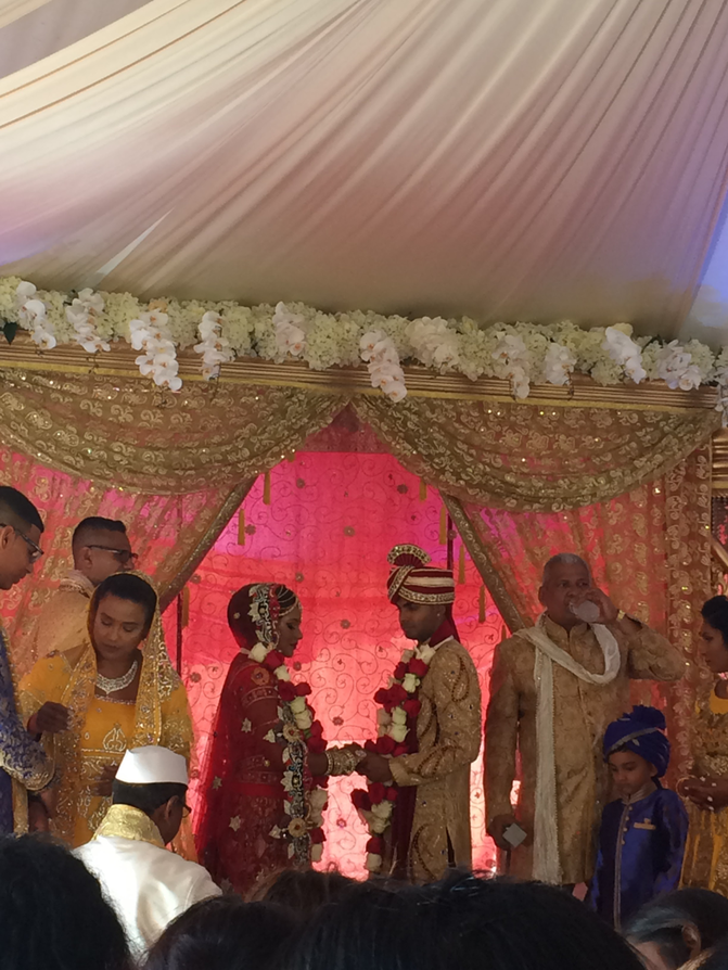 A photo of the Hindu wedding that the author attended. In the center is the bride and groom wearing traditional Indian garb for a Hindu wedding. To the left, the bride is wearing a saree and a veil. To the right, the groom is wearing a kurta and a safa. To the right of the groom, the groom’s father is wearing a more traditional kurta, with a scarf, but without the safa.
