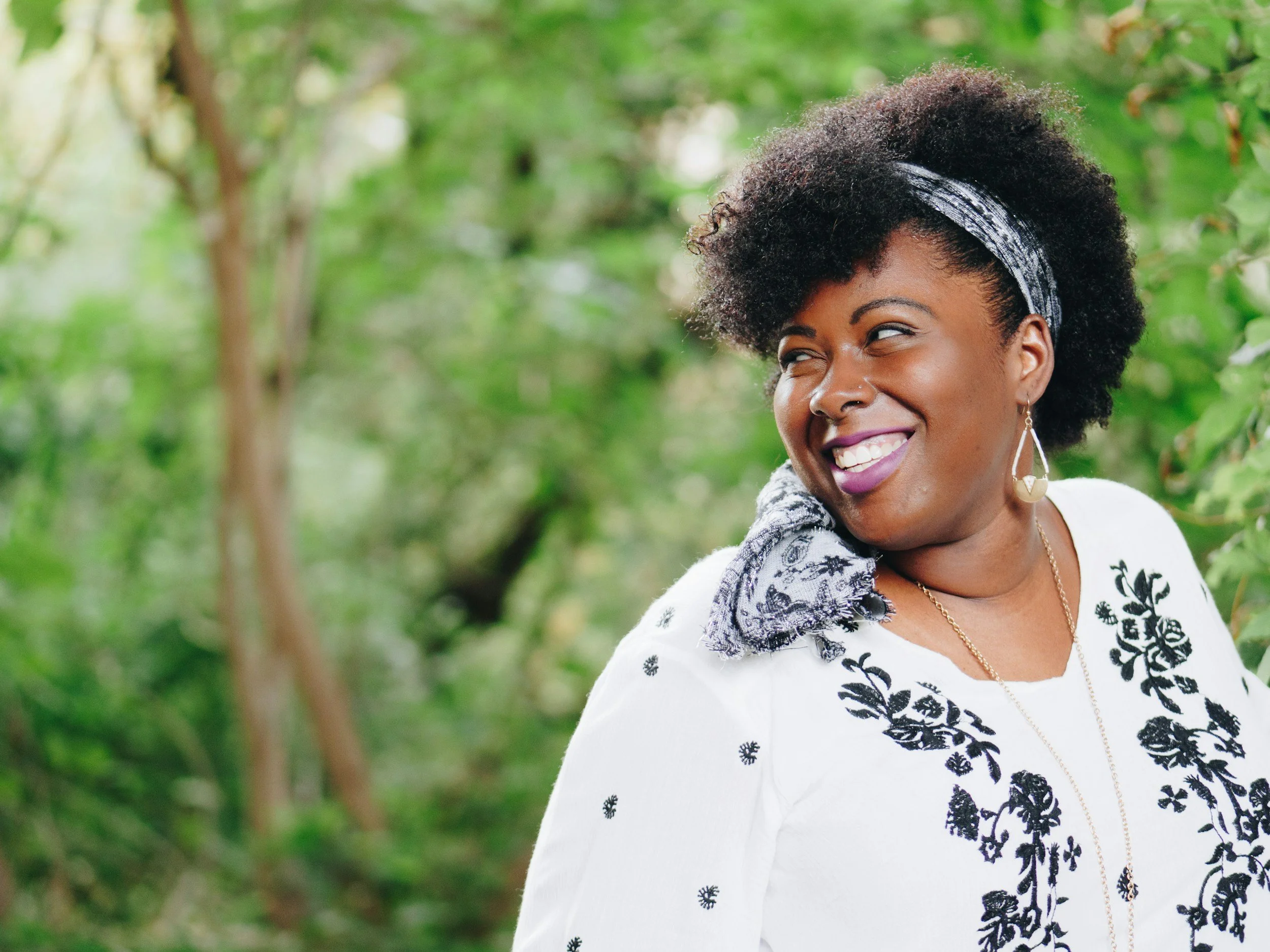 A woman with curly black hair wearing a patterned headband, white blouse with black floral embroidery, and hoop earrings, smiling and looking to her right outdoors amidst green foliage.