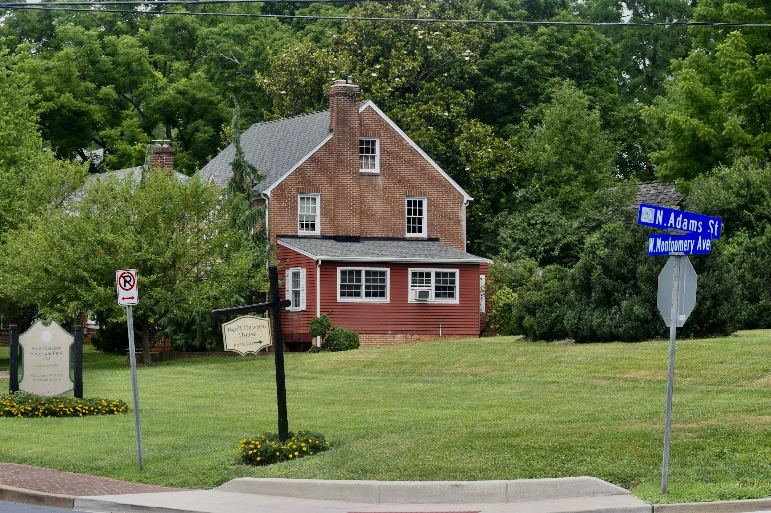 A brick house with red siding on a grassy area, surrounded by trees, with street signs for N. Adams St and W. Montgomery Ave, and a sign for Beall-Dawson House historic park.