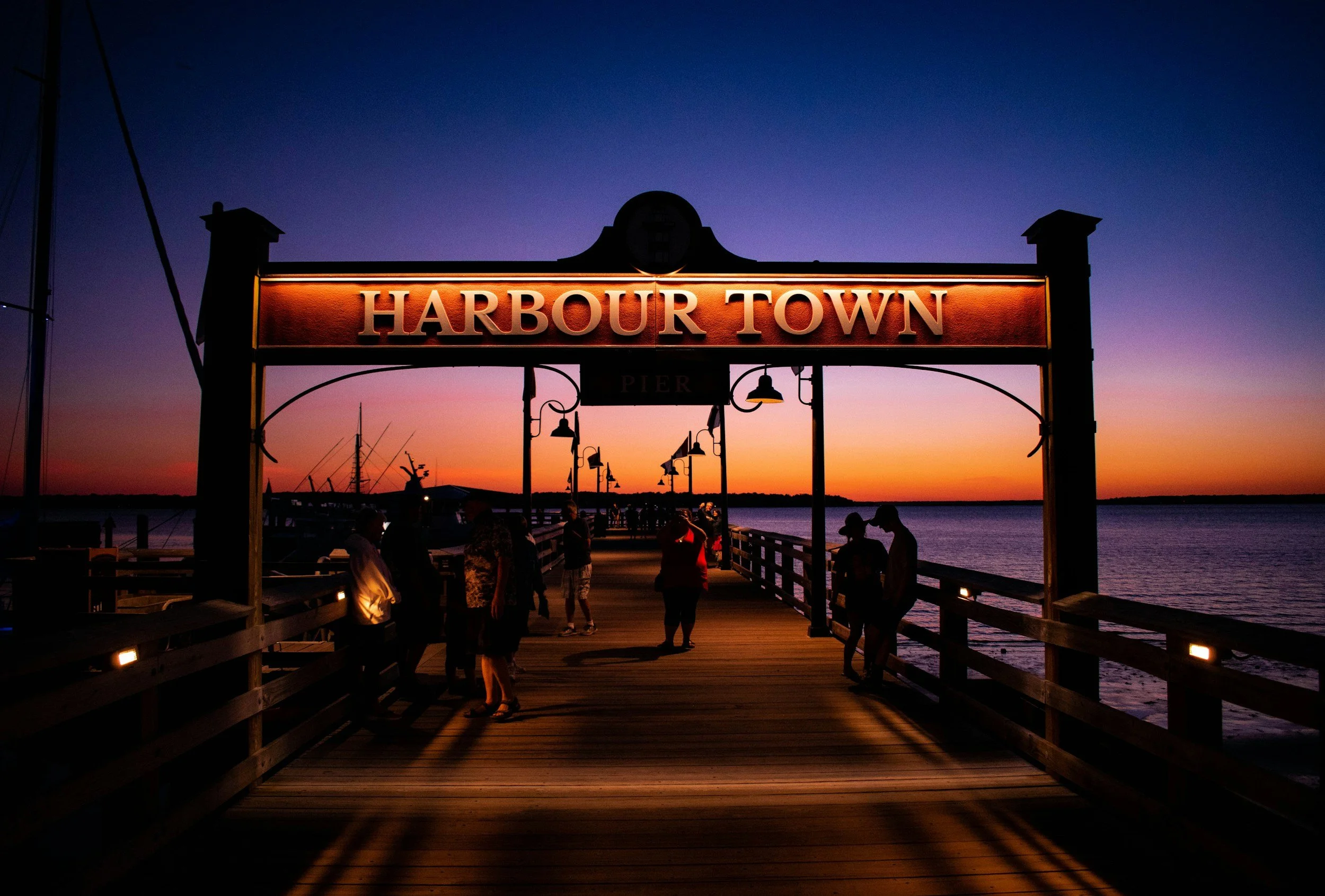 A pier at sunset with a sign reading 'Harbour Town', silhouetted people walking, and boats docked nearby under a colorful sky.