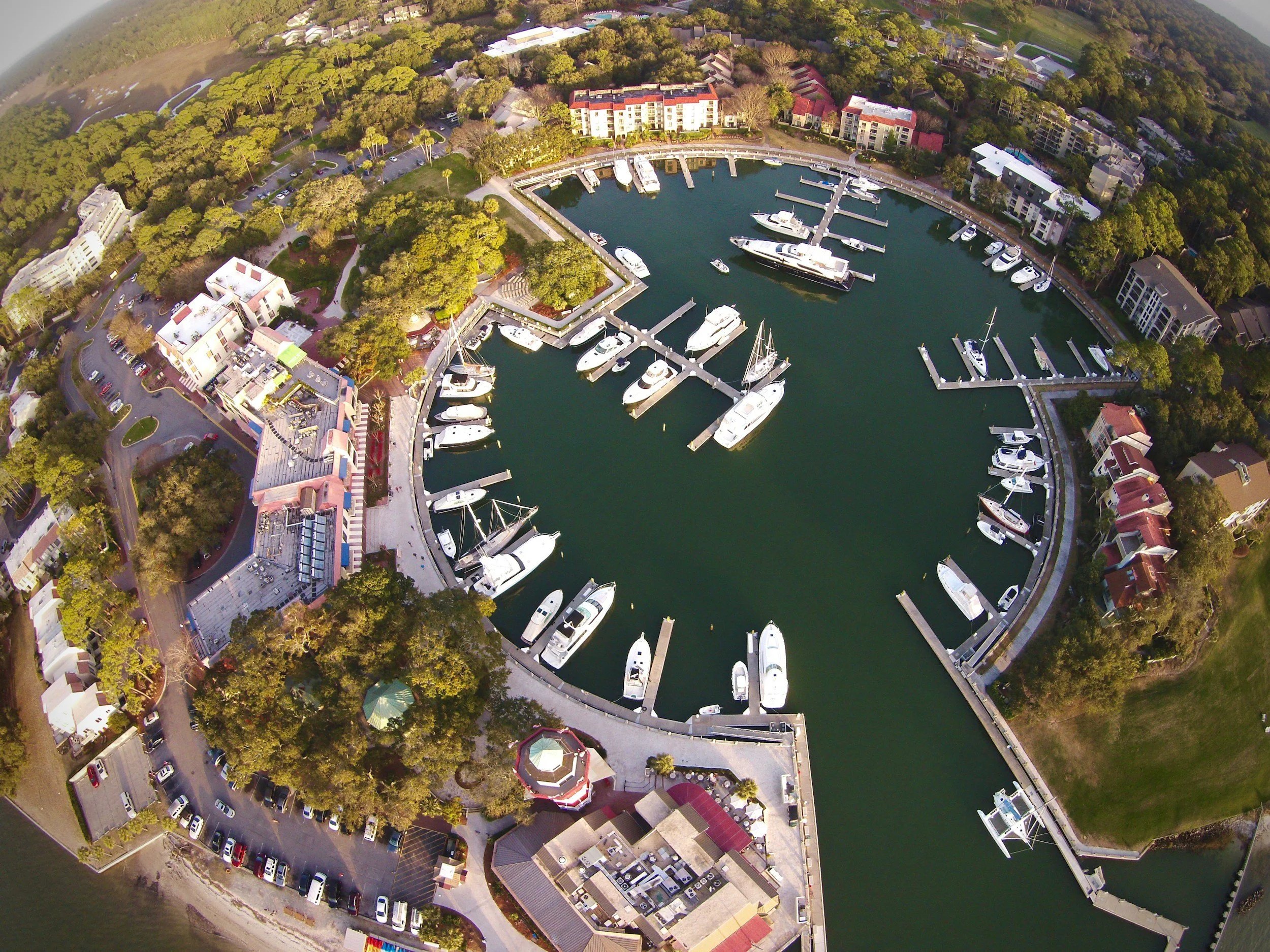 An aerial view of a marina with boats docked in the water, surrounded by residential buildings, trees, and parking lots.