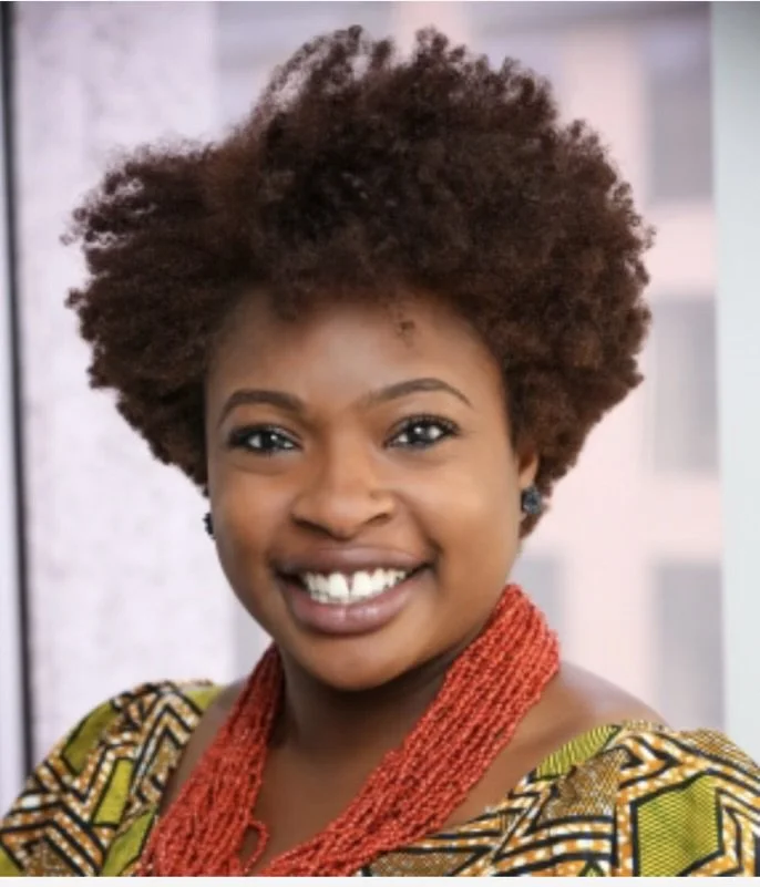 A woman with short, curly, brown hair smiling, wearing a colorful patterned top and a red beaded necklace, indoors.