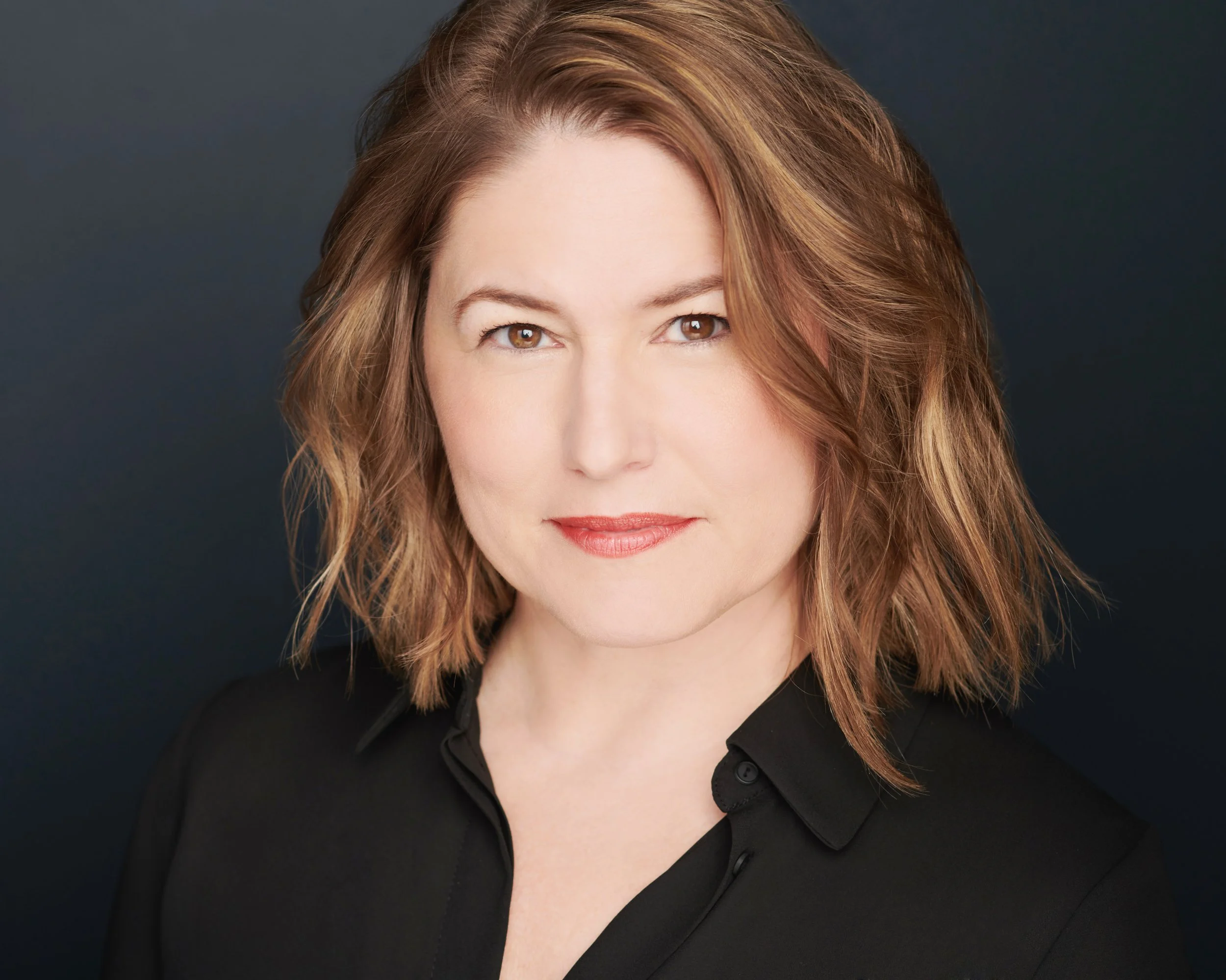 A woman with shoulder-length auburn hair, wearing a black top, looking at the camera with a subtle smile against a dark background.