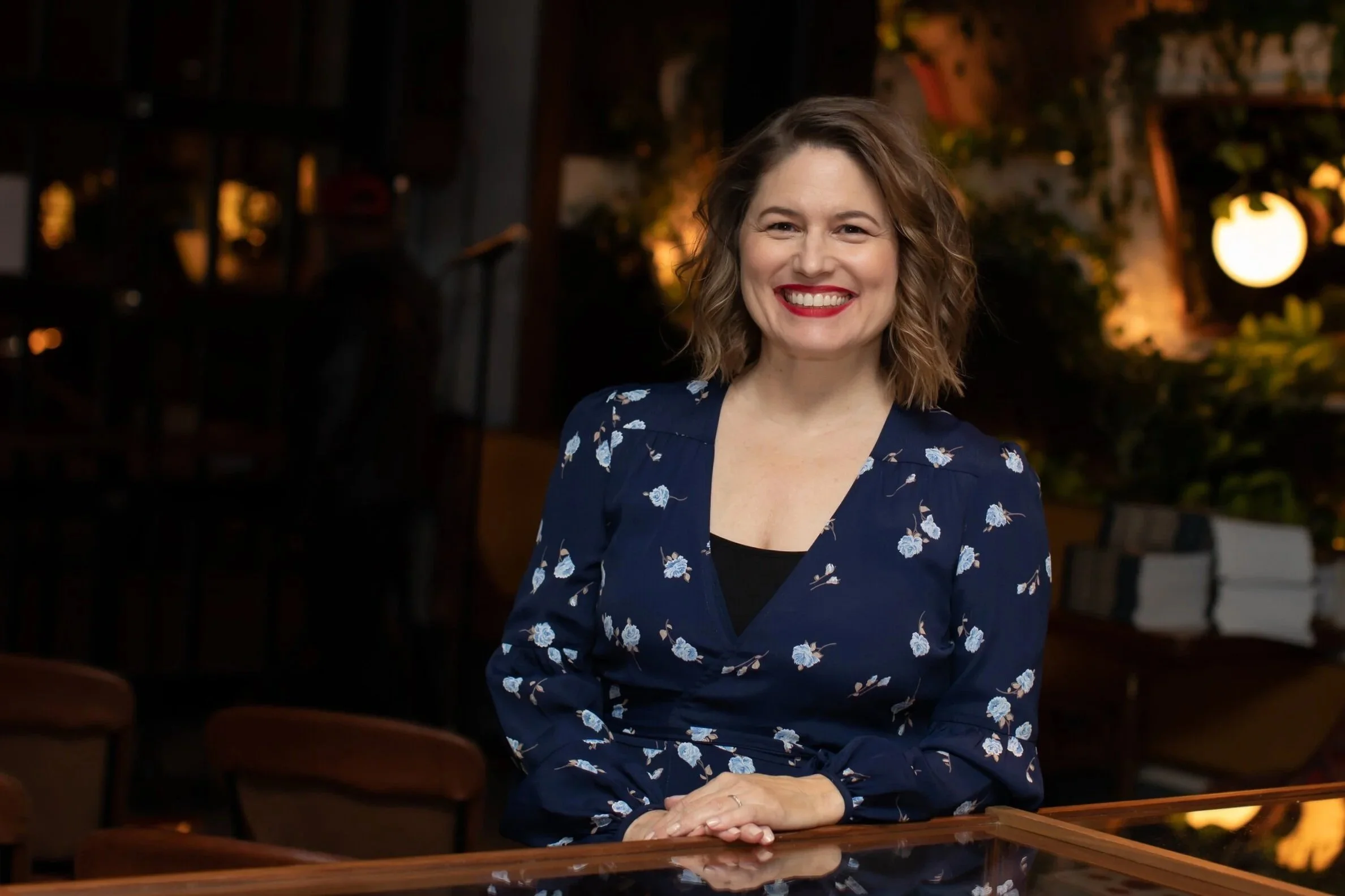 A woman with shoulder-length curly hair, wearing a navy blue floral dress, smiling while sitting at a bar in a warmly lit, cozy indoor setting.