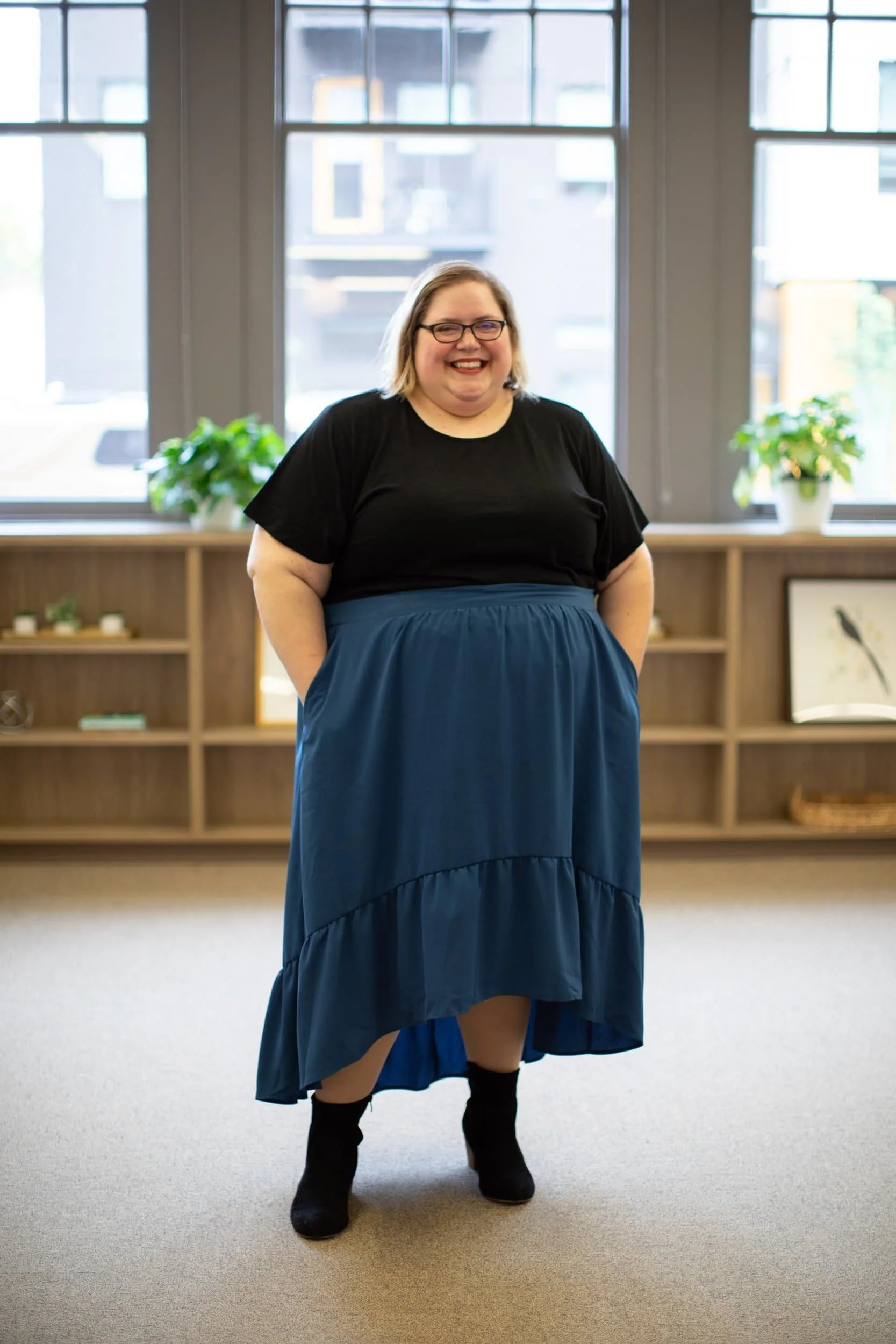 A woman smiling, wearing glasses, a black top, a long blue skirt, and black shoes, standing indoors in front of windows with potted plants on a shelf behind her.