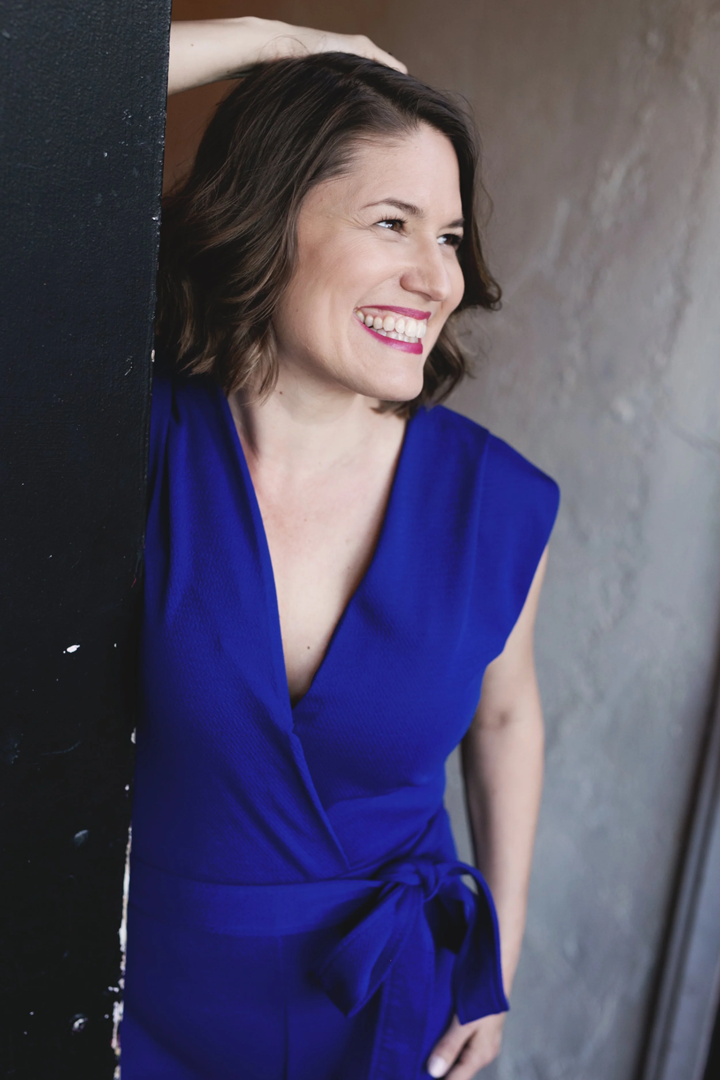 A smiling woman with short brown hair, wearing a blue sleeveless top, standing against a gray wall, with one arm resting on a black textured surface.