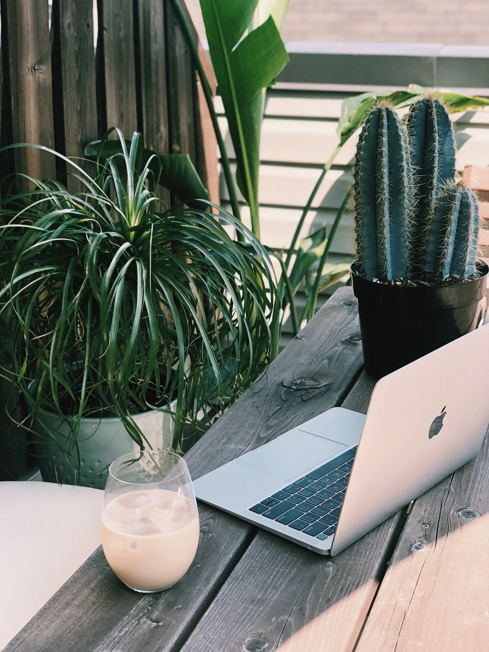 Wooden table with a glass of iced coffee and an apple laptop with green plants behind it.
