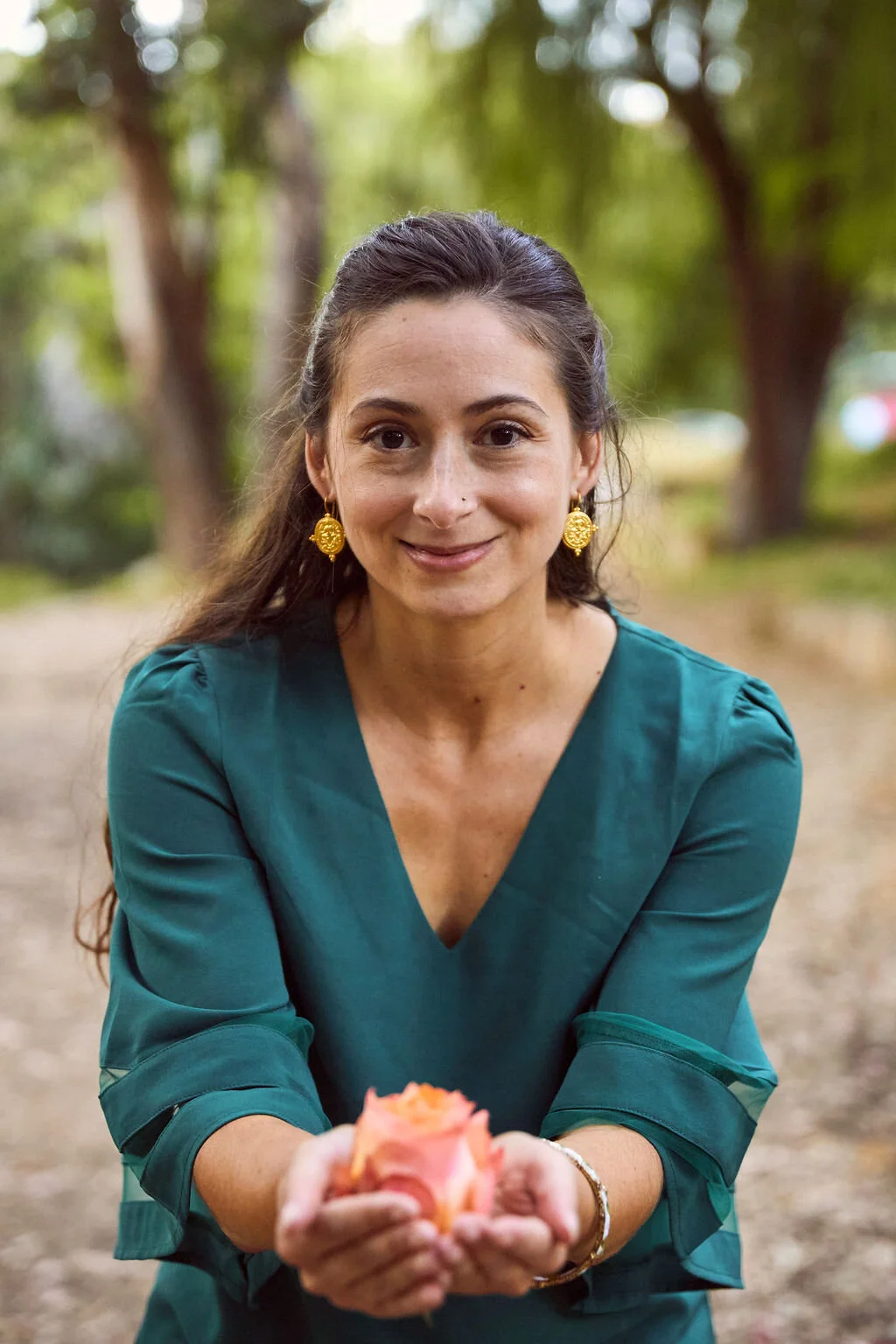 A woman with long dark hair and gold earrings smiling outdoors, holding flowers in her hands, with trees and a blurred background.