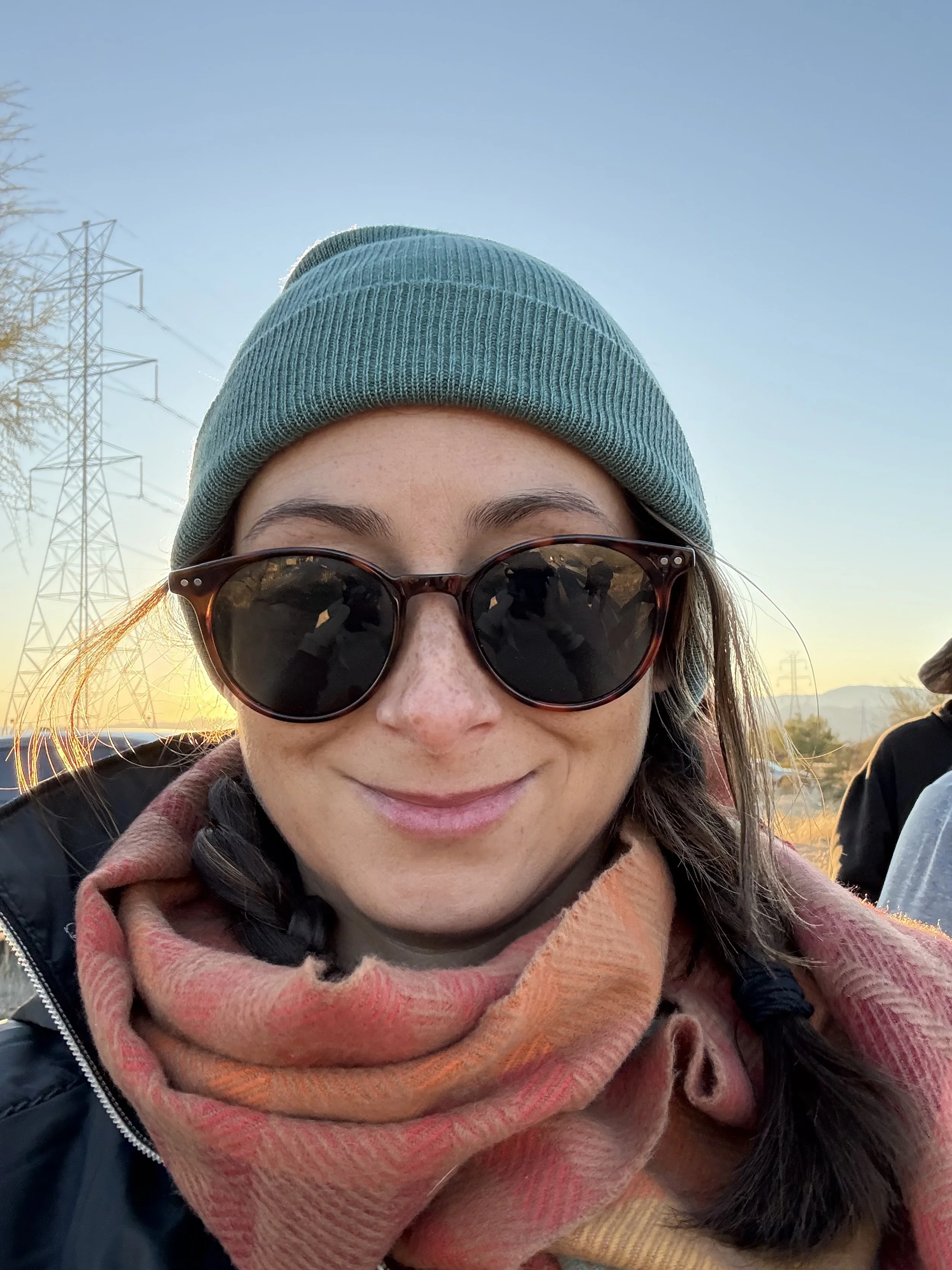 A woman wearing sunglasses, a teal beanie, and a multicolored scarf, smiling outdoors during sunset.