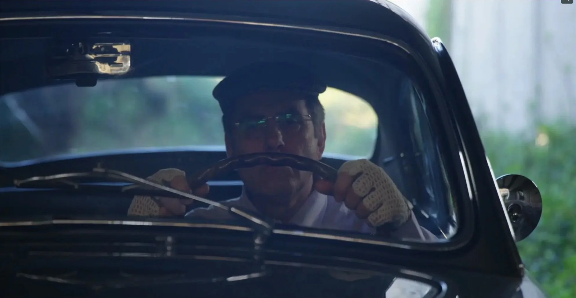 A person wearing glasses and a cap, sitting in a vintage car, holding the steering wheel from behind the dashboard.