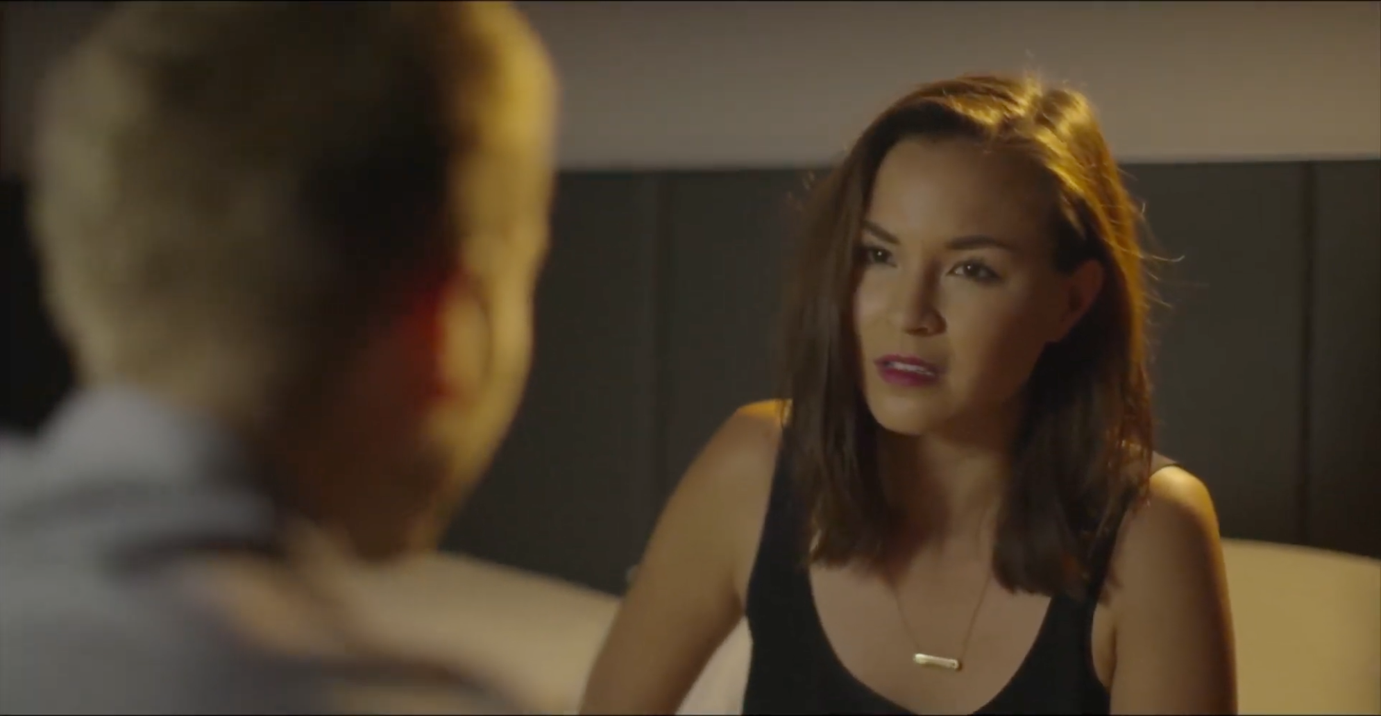 A woman with shoulder-length brown hair and a black top talking to a man whose face is not visible, in an indoor setting with a dark wall background.