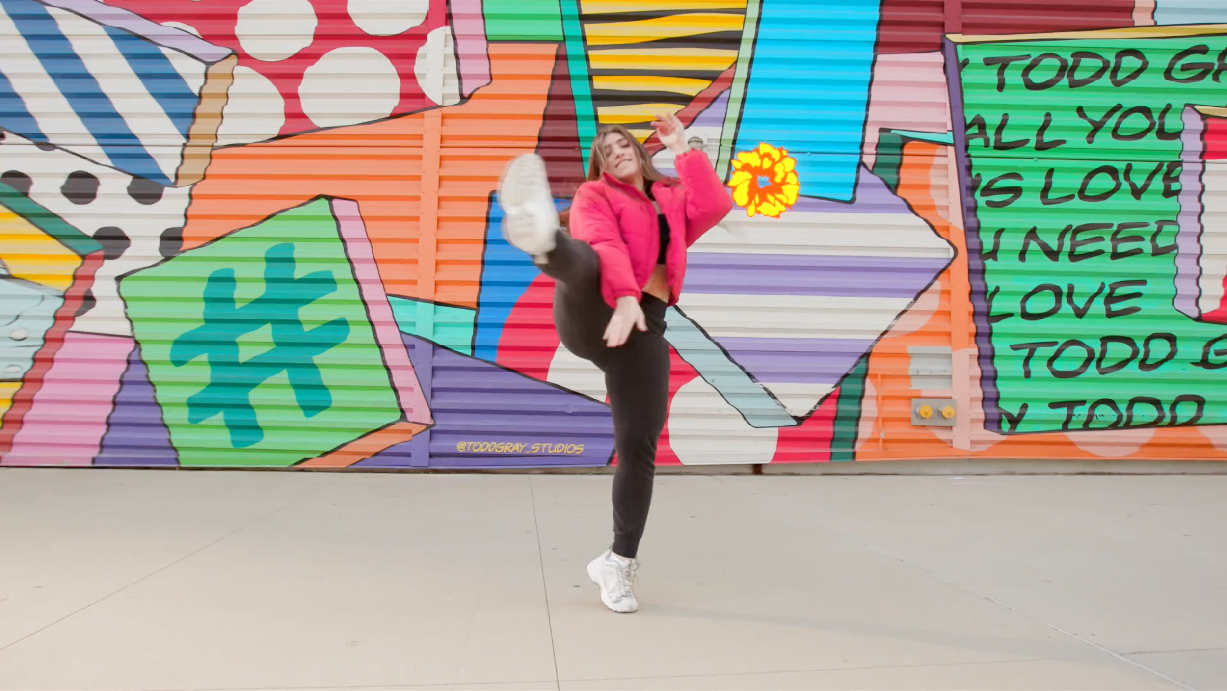 Woman in pink jacket and black pants performing a dance move in front of colorful graffiti art wall