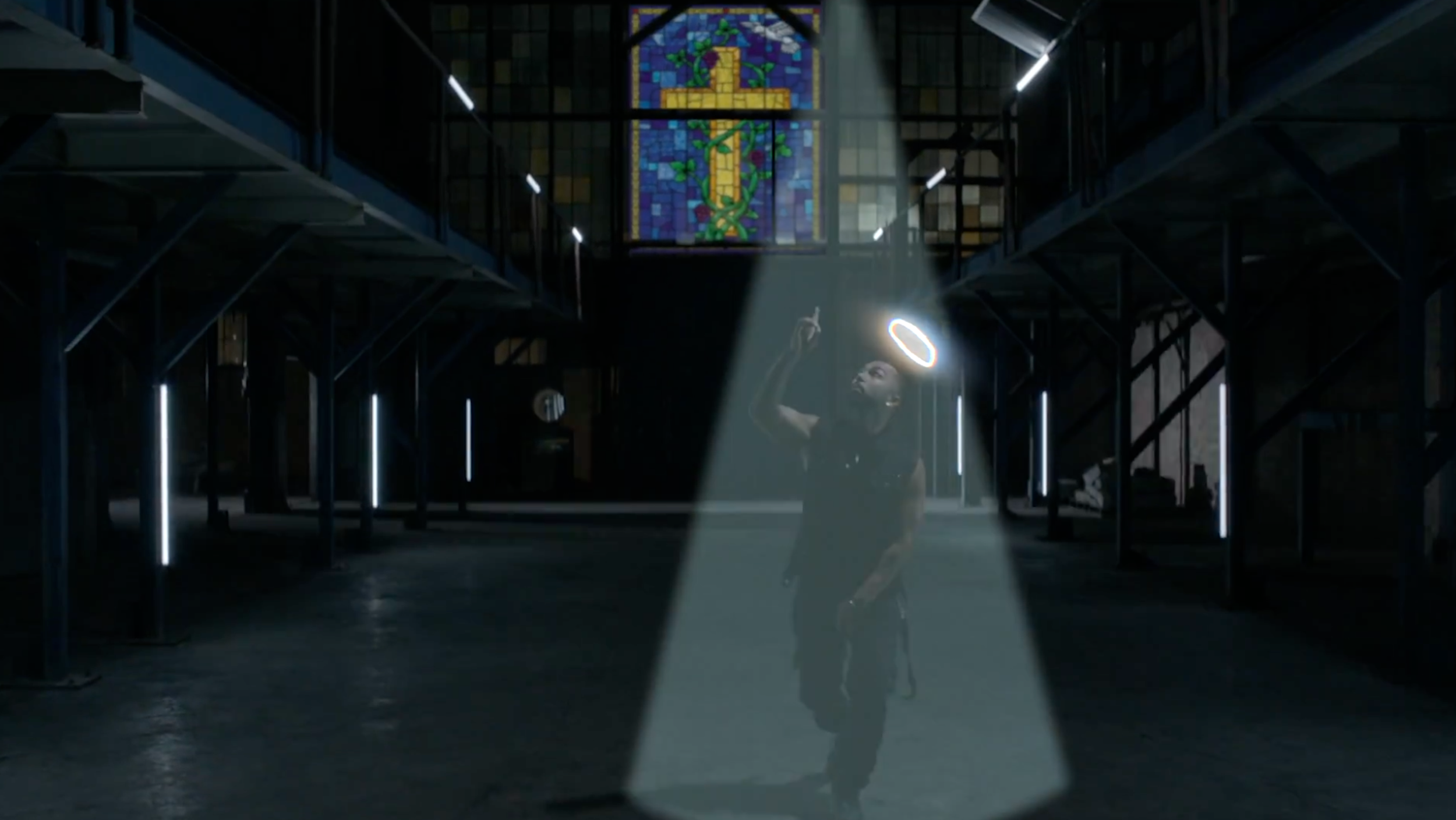 A man dressed in black with a halo-like ring floating above his head, standing under a spotlight casting a shadow on the floor inside a dark, spacious building with a staircase on each side and a stained glass window featuring a cross at the back.