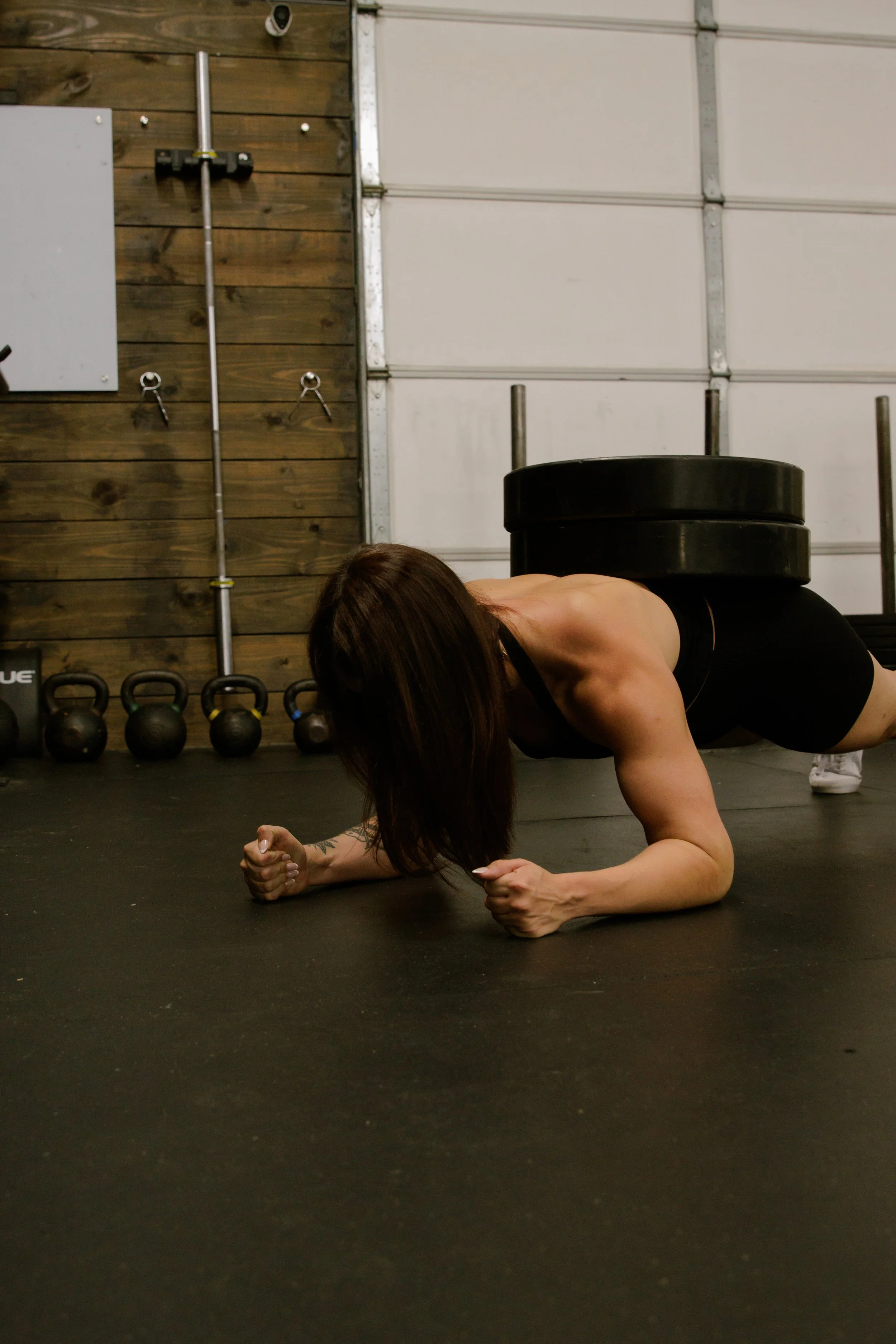 A woman doing a plank exercise with a weight plate on her back in a gym.