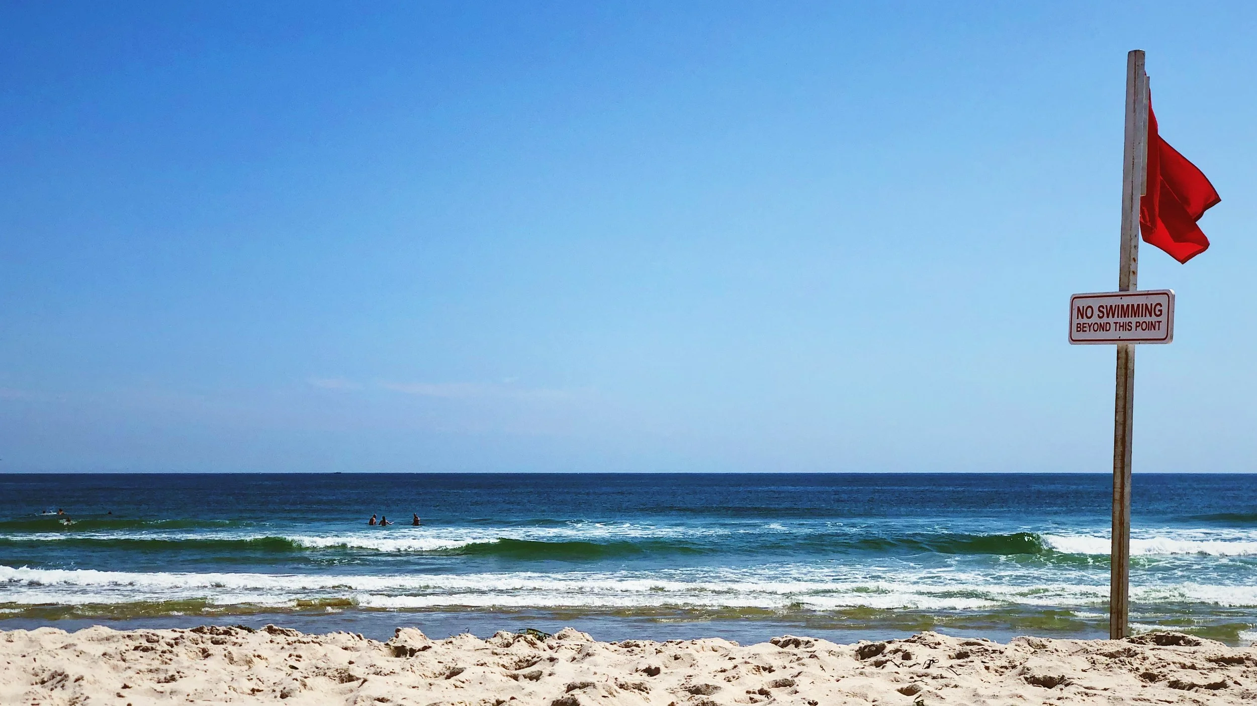 Ocean waves breaking upon then beach with a "no swimming beyond this point" sign.