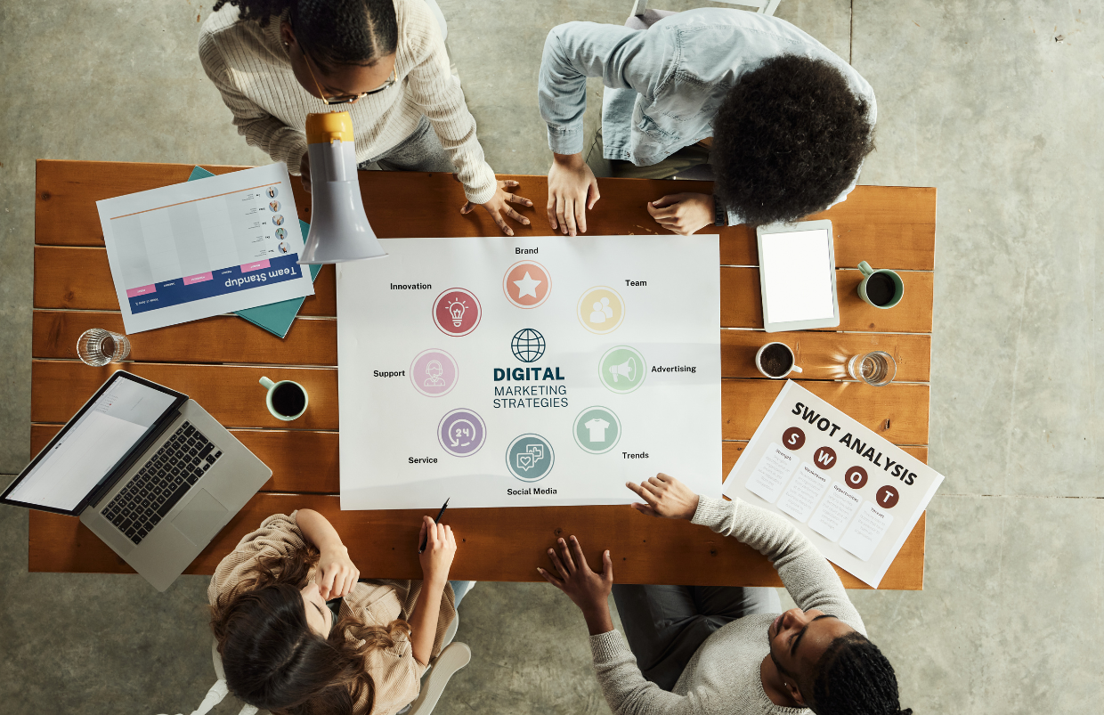 Top view of diverse team collaborating on digital marketing strategies around a wooden table, with a laptop, SWOT analysis document, and brainstorming materials.