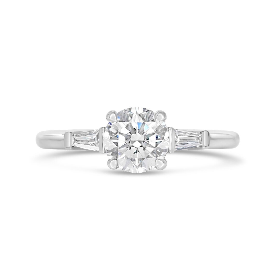 A finely balanced trilogy with tapered baguette diamonds brings a subtle geometric influence to this design while still maintaining the classic softness of the centre round brilliant diamond.