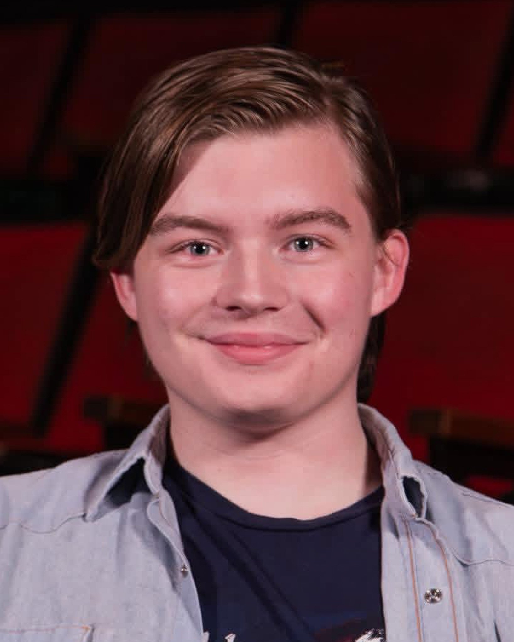 Close-up of a young person's face with short brown hair, blue eyes, smiling, wearing a black shirt and a grey button-up shirt, against a blurry red and black background.