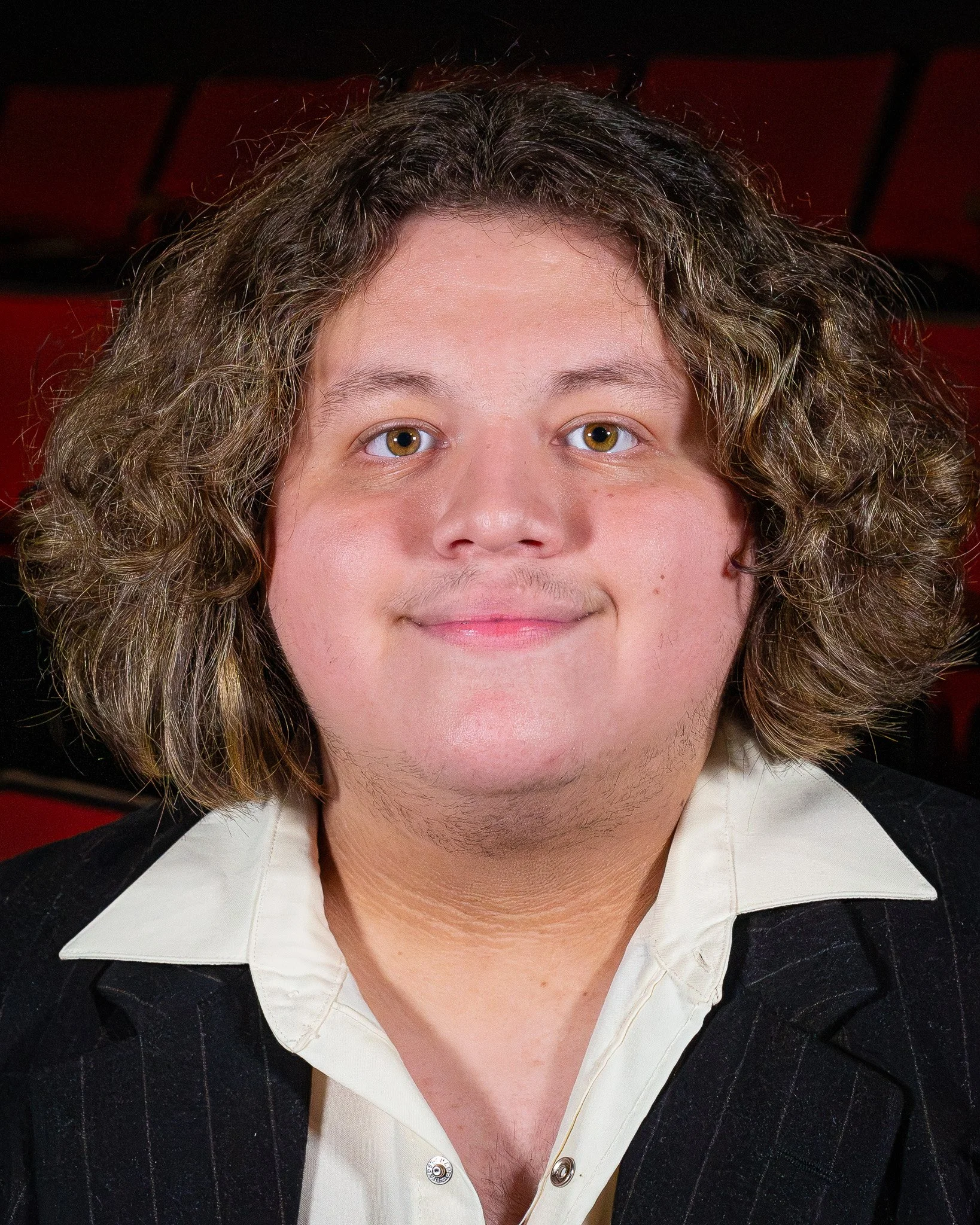 Close-up of a person's face with curly hair, light skin, and hazel eyes, wearing a white shirt and a dark, pinstriped blazer, sitting in a theater with red seats in the background.