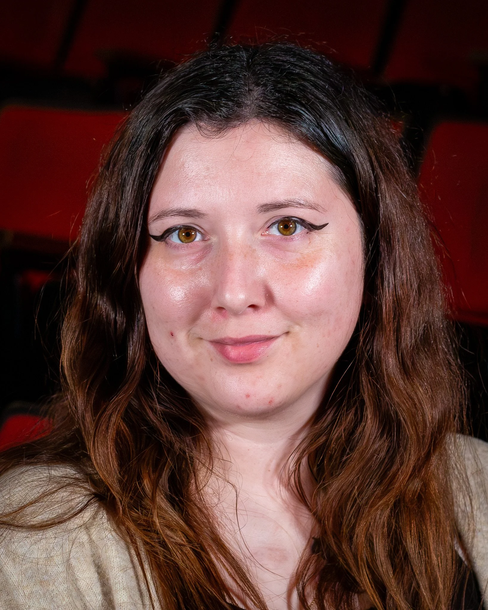 Close-up of a woman with brown hair and brown eyes, wearing black eyeliner, against a dark red background.