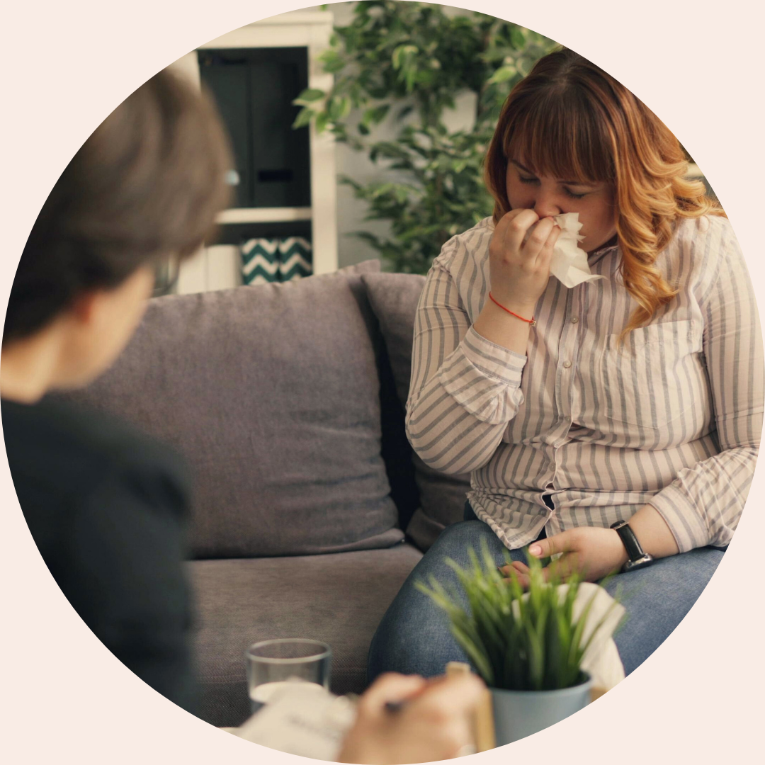 A woman sitting on a couch holds a tissue to her face while appearing tearful during a counselling session, with a therapist seated opposite taking notes in a calm, supportive setting.