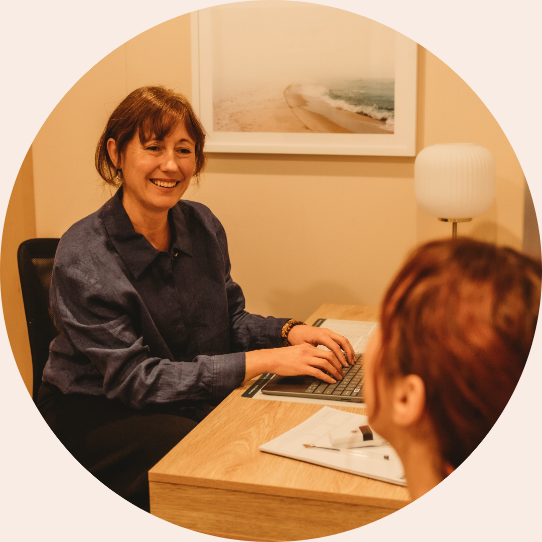 A female therapist sits at a desk smiling warmly while speaking with a client, with a notebook and laptop on the table in a calm, softly lit counselling office.